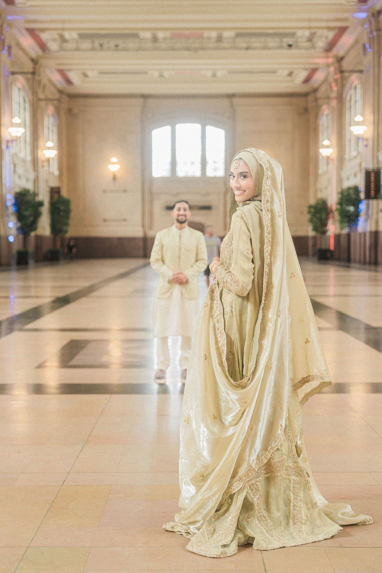 A bride in a gold hijab and gown stands elegantly in a grand hall as her groom approaches in matching attire.