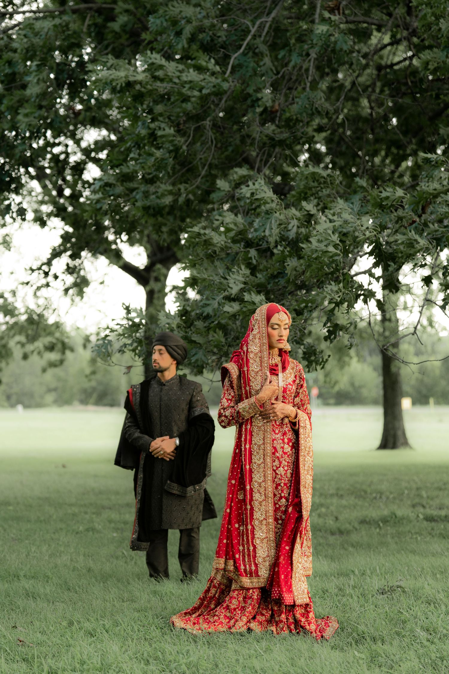 South Asian bride in red bridal lehenga and groom in dark sherwani standing under a tree in a lush green park.