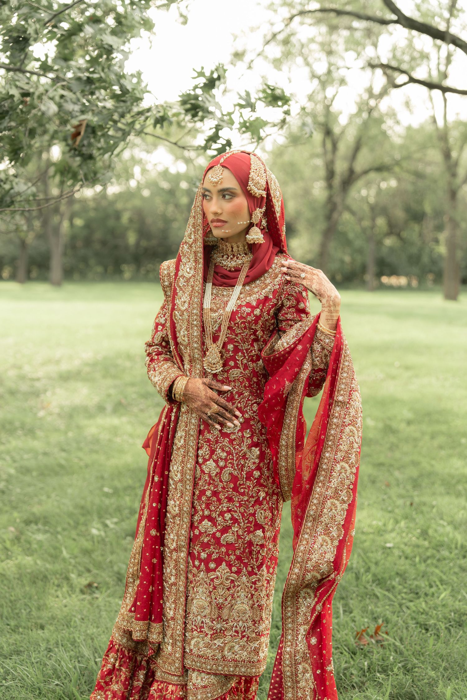 A bride in a red and gold embroidered lehenga with dupatta stands gracefully outdoors among green trees.