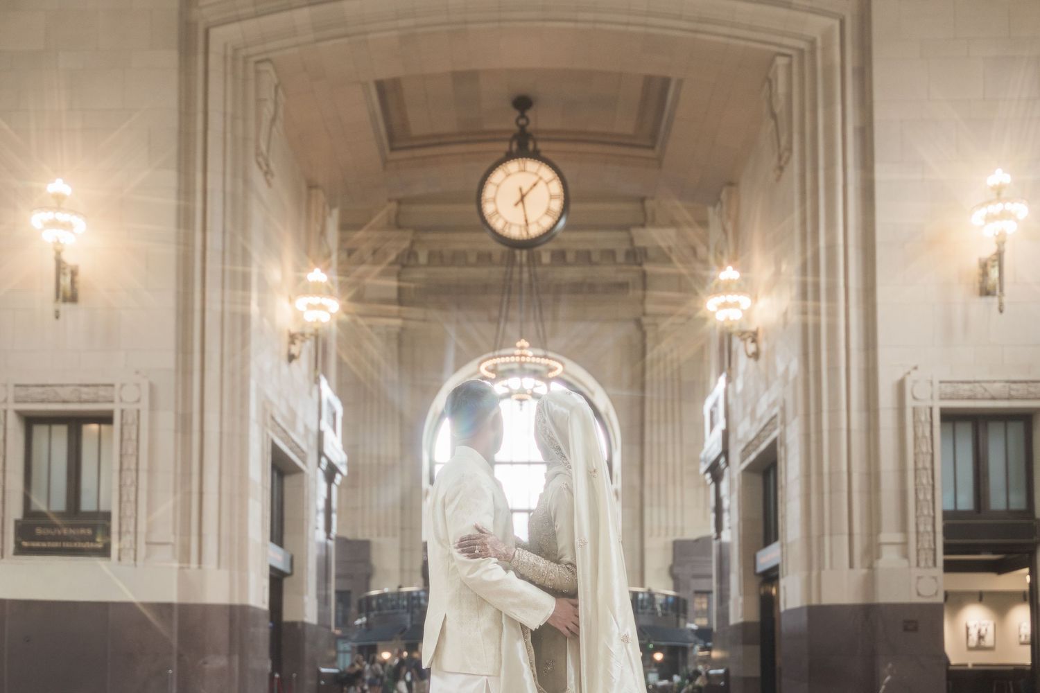 Elegant wedding couple embracing in a grand hall with ornate architecture, chandeliers, and a classic clock overhead.