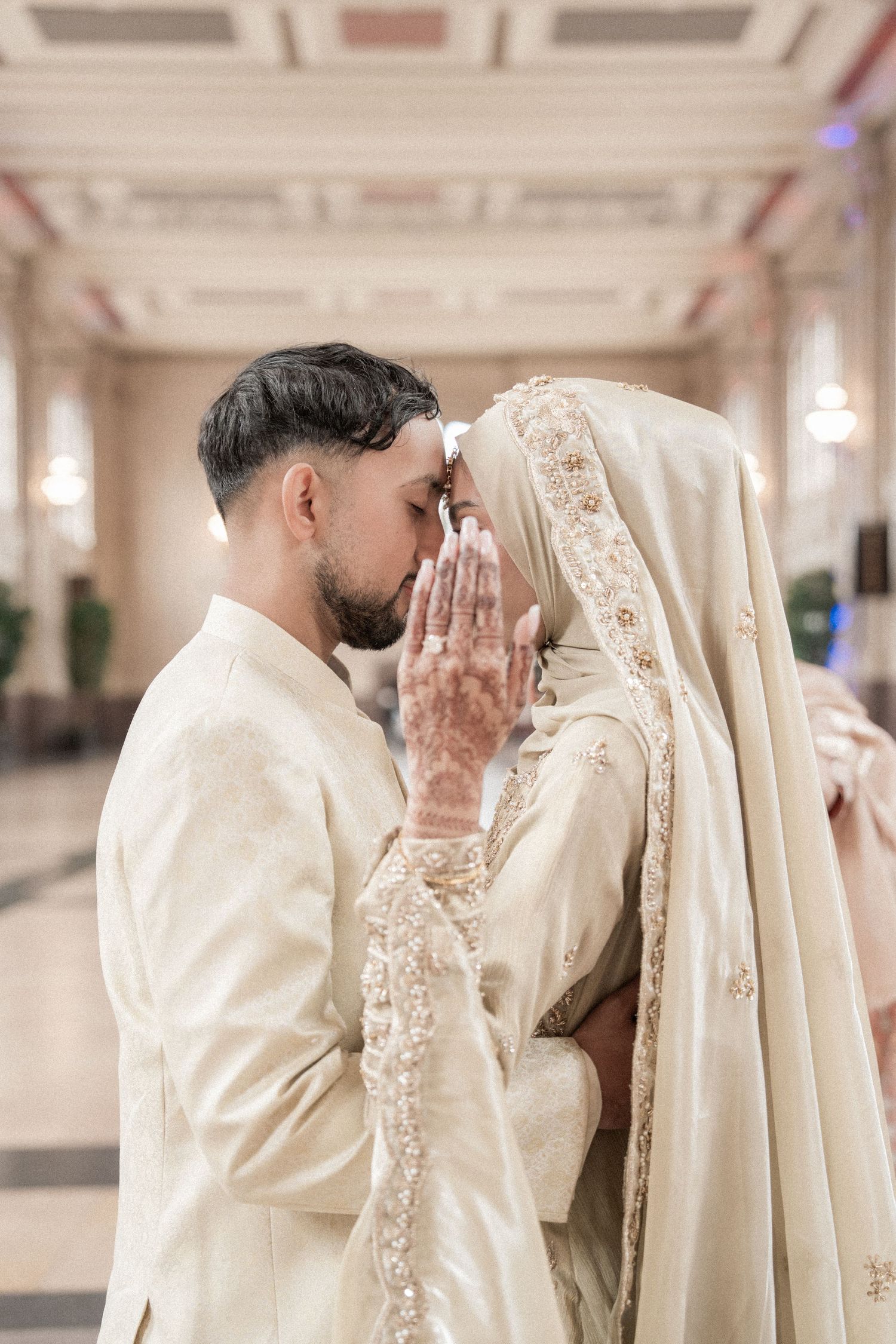South Asian bride and groom in ivory wedding attire share an intimate moment, bride wearing henna and hijab.