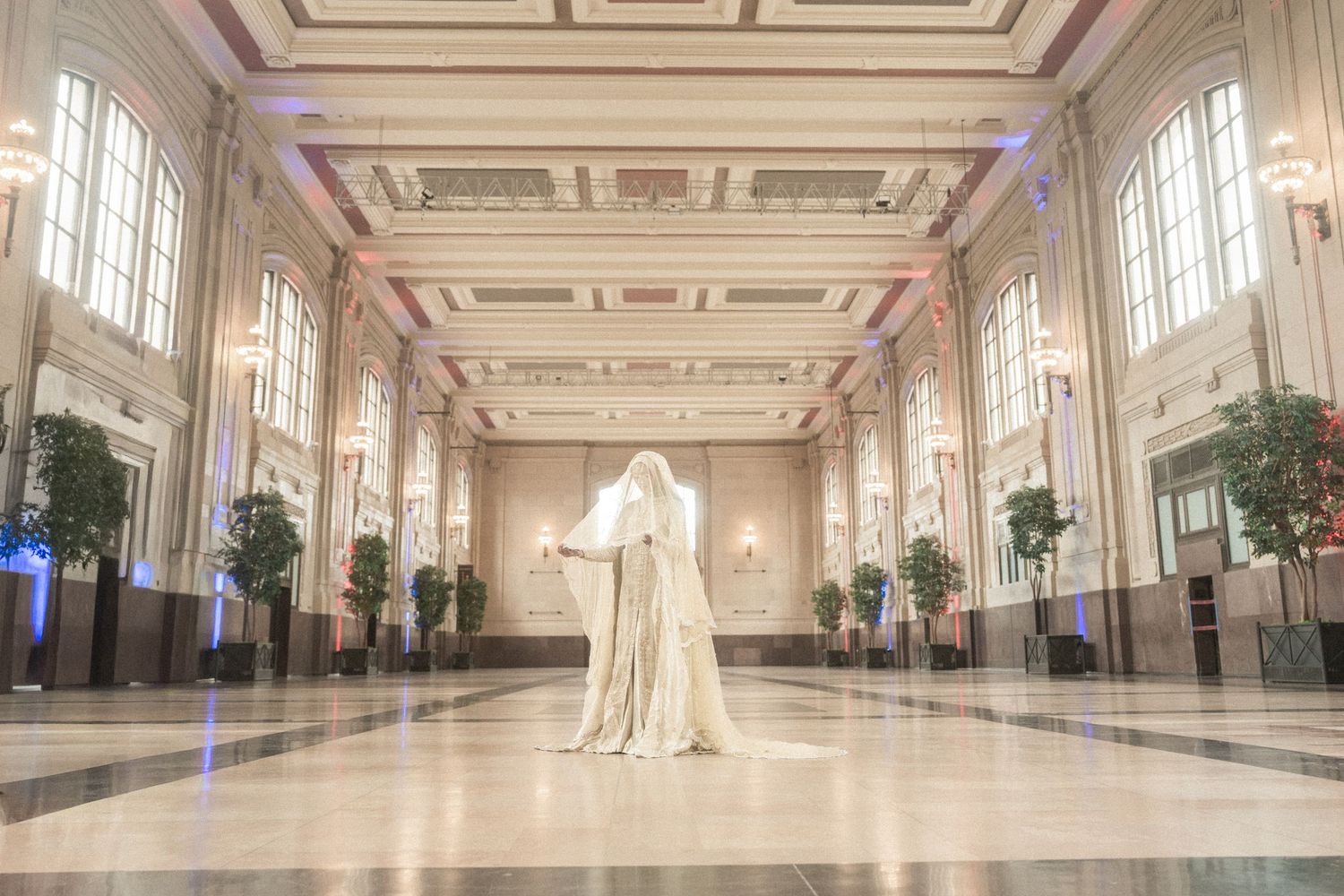 A robed figure stands in an elegant grand hall with ornate ceilings, marble floors, and decorative trees lining the walls.