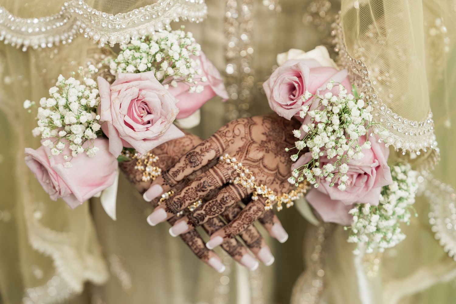 Two henna-adorned hands holding pink roses and baby's breath bouquets at a traditional South Asian wedding ceremony.