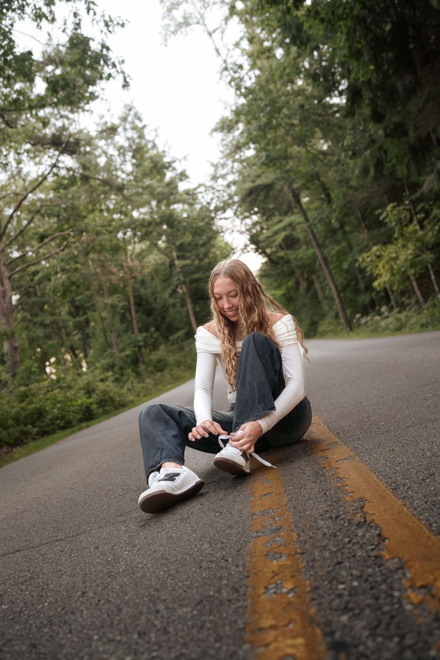 Person in casual attire sitting on an asphalt road with double yellow lines and trees in background.