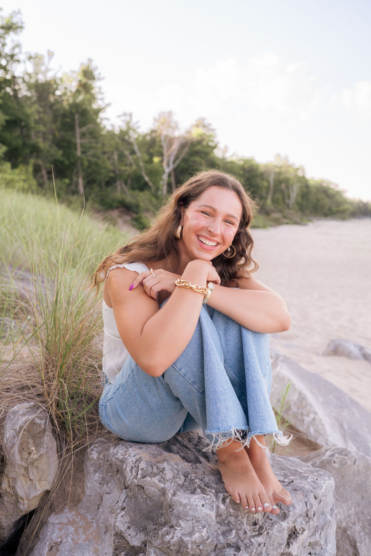 A casual beach portrait shows someone sitting on a rock wearing jeans and a white top against a coastal backdrop.