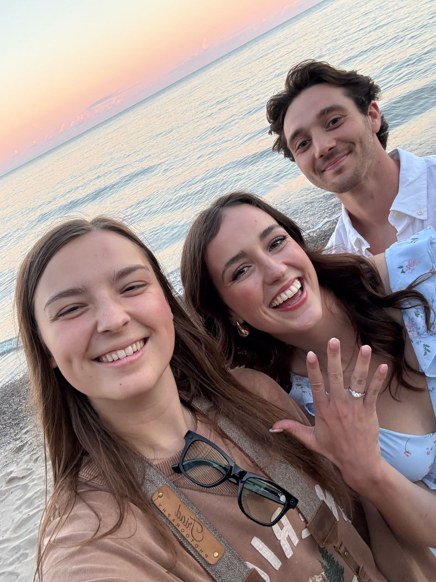 A sunset beach selfie with three people smiling, one showing off a ring.