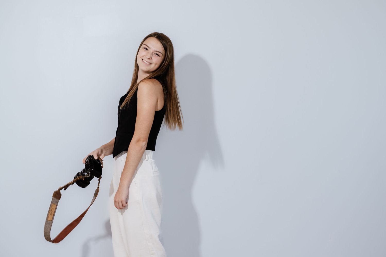 Professional wearing black top and white pants sits casually against white background in studio setting.