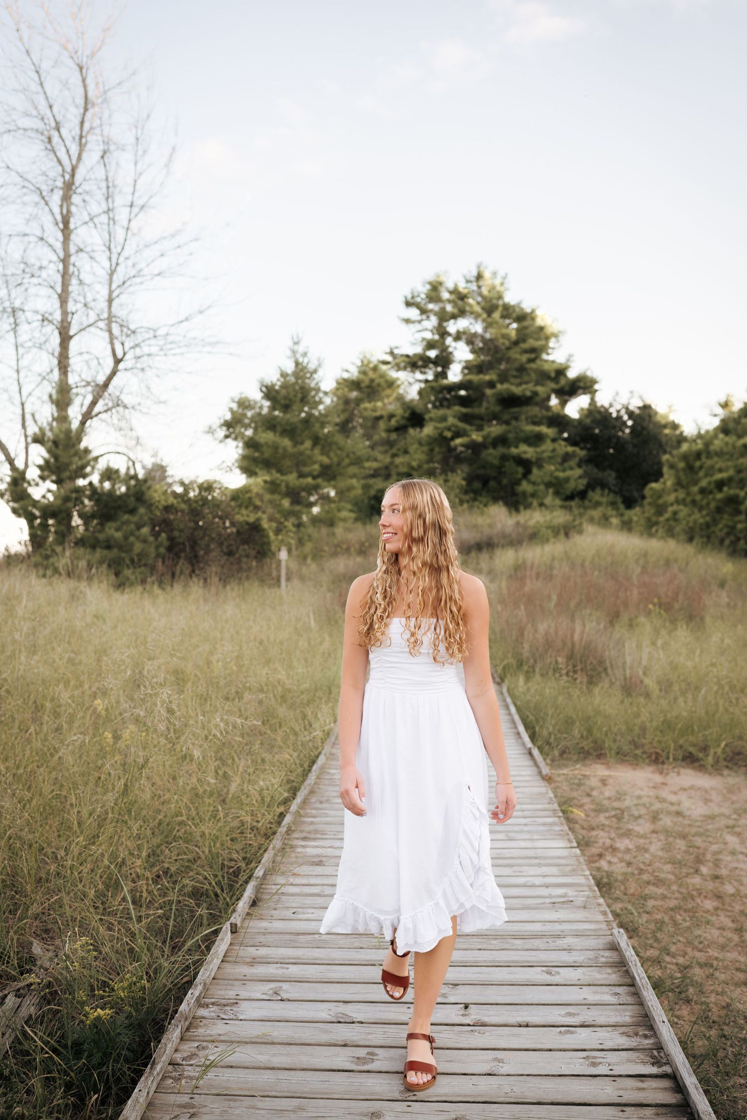 Person in flowing white dress walking along wooden boardwalk through natural grassland.