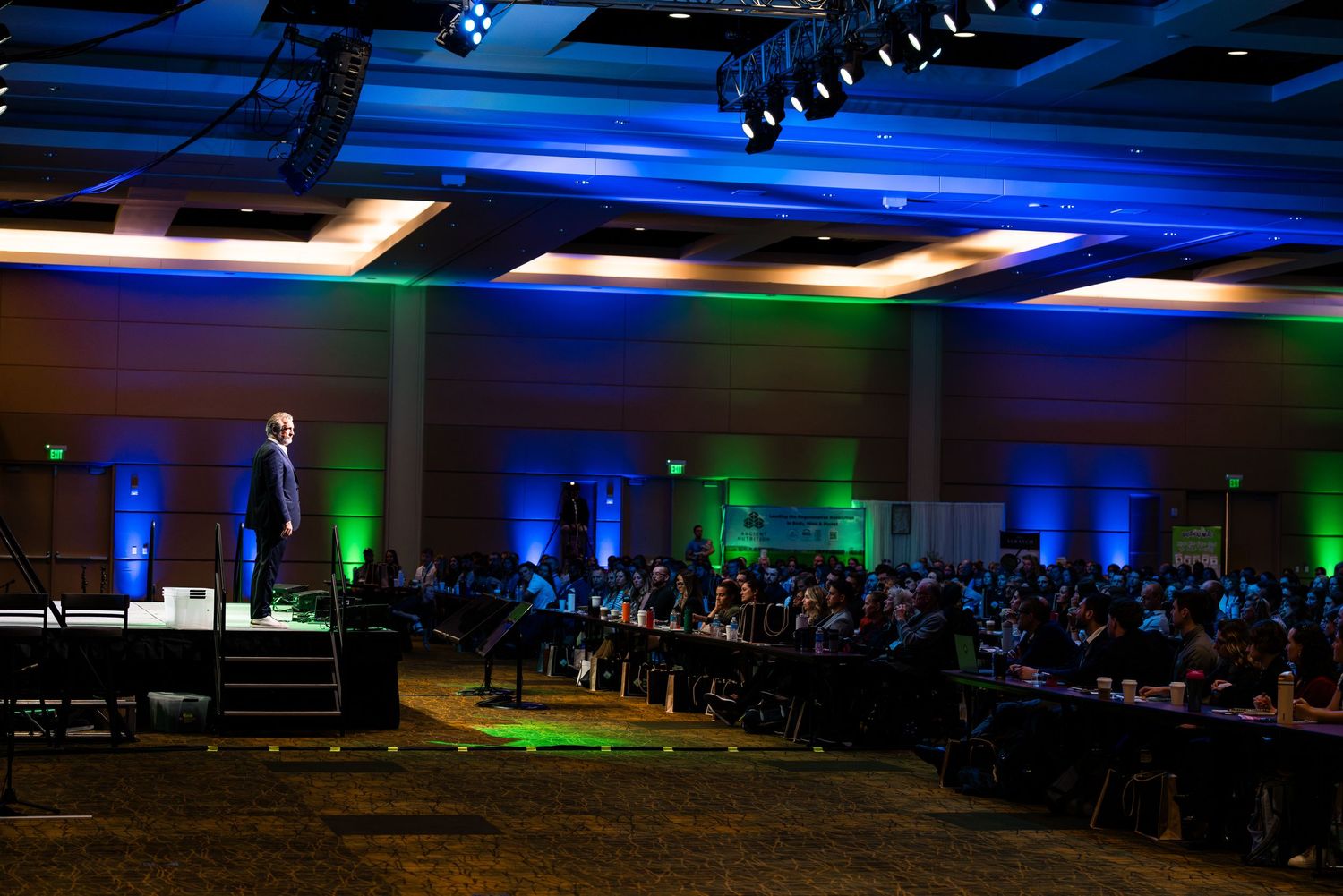 Wide conference hall view with blue ambient lighting and rows of attendees.