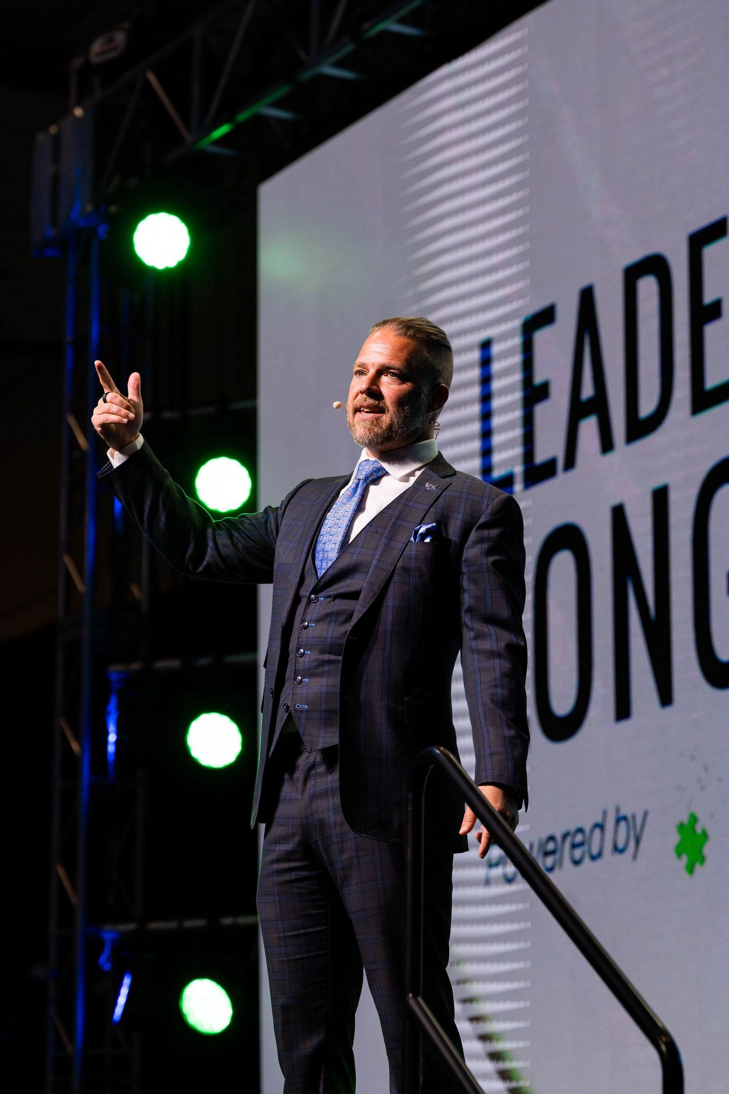 Speaker in a dark suit gestures while presenting on stage with branded backdrop.