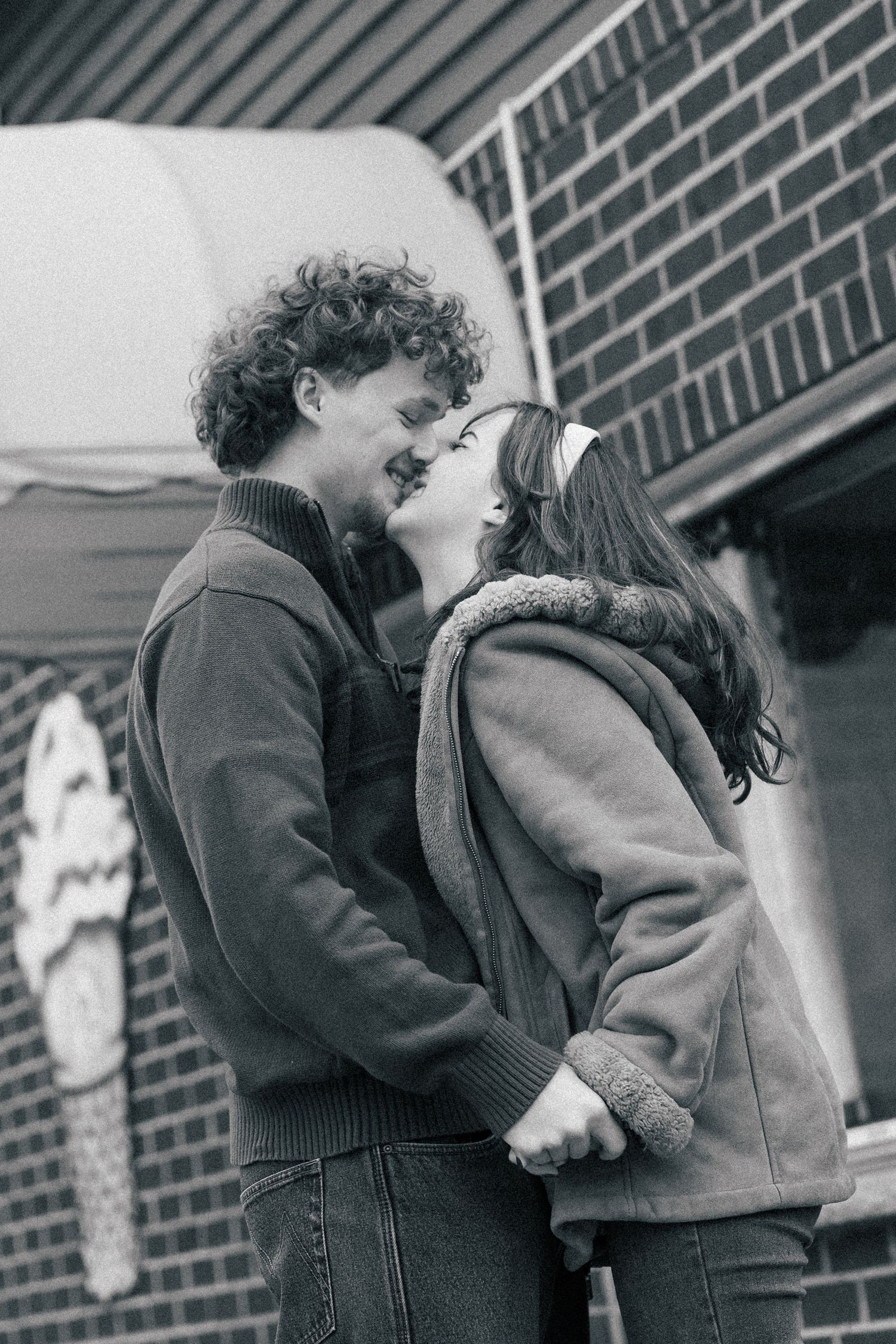Black and white photo of a young couple holding hands and sharing a romantic moment outside a brick building.