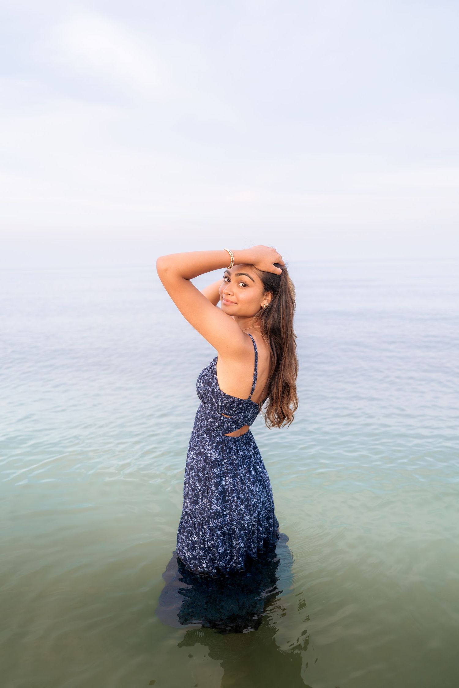 Woman in blue floral dress poses artistically in shallow water during sunset.