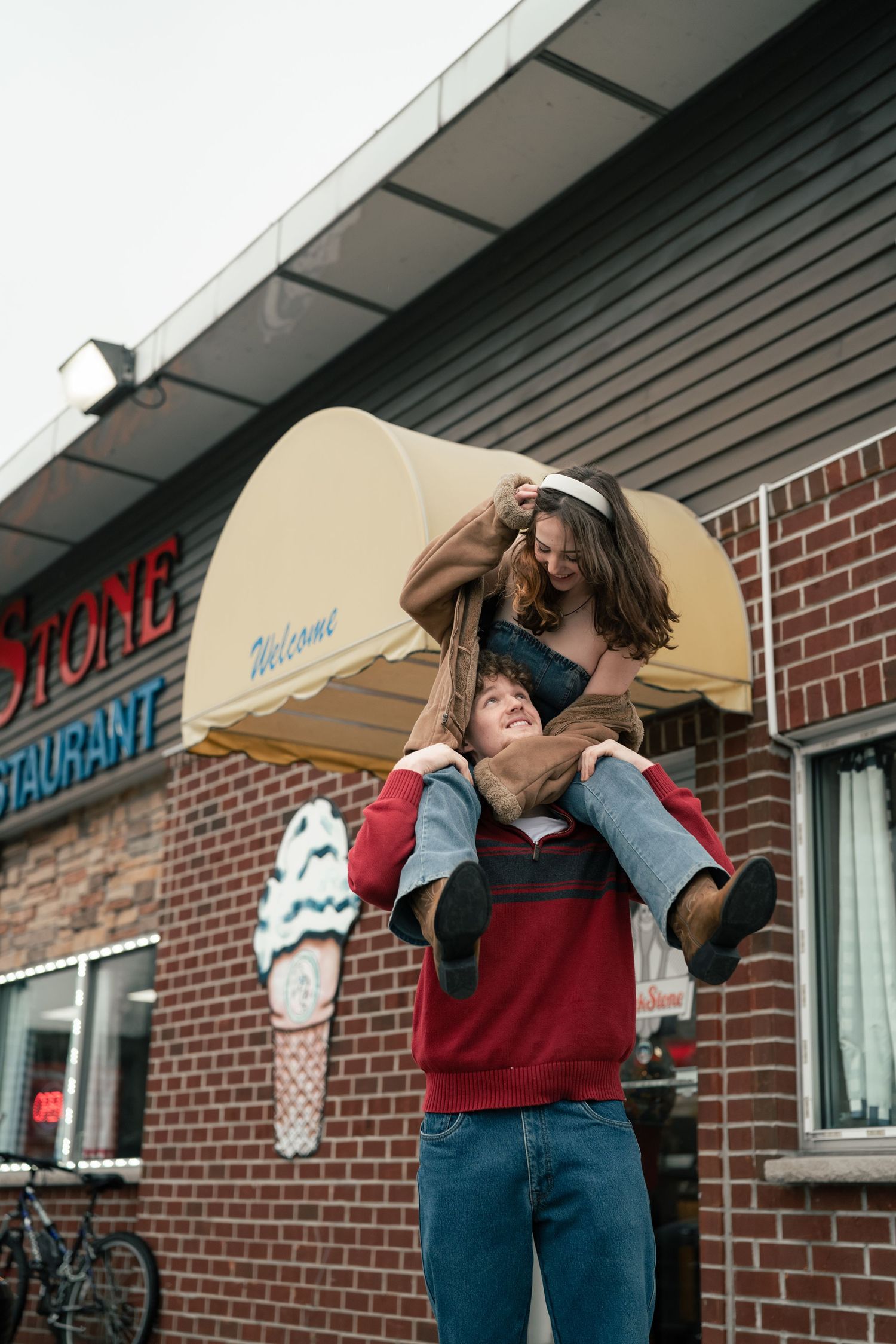 A young couple laughing outside a brick restaurant, man lifting woman on his shoulders near a welcome awning.