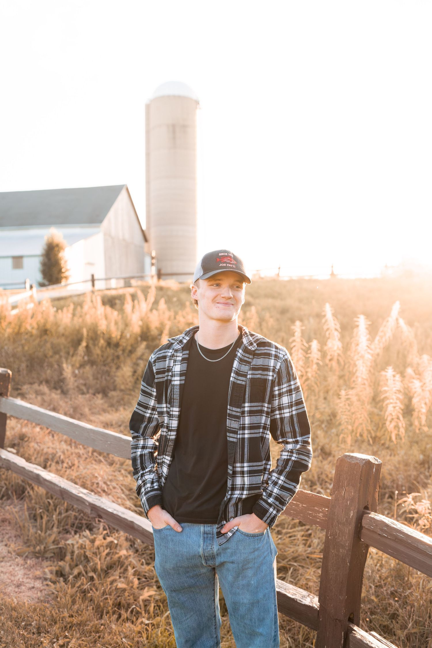 A casual portrait of someone in plaid and jeans standing by a rustic barn and silo.
