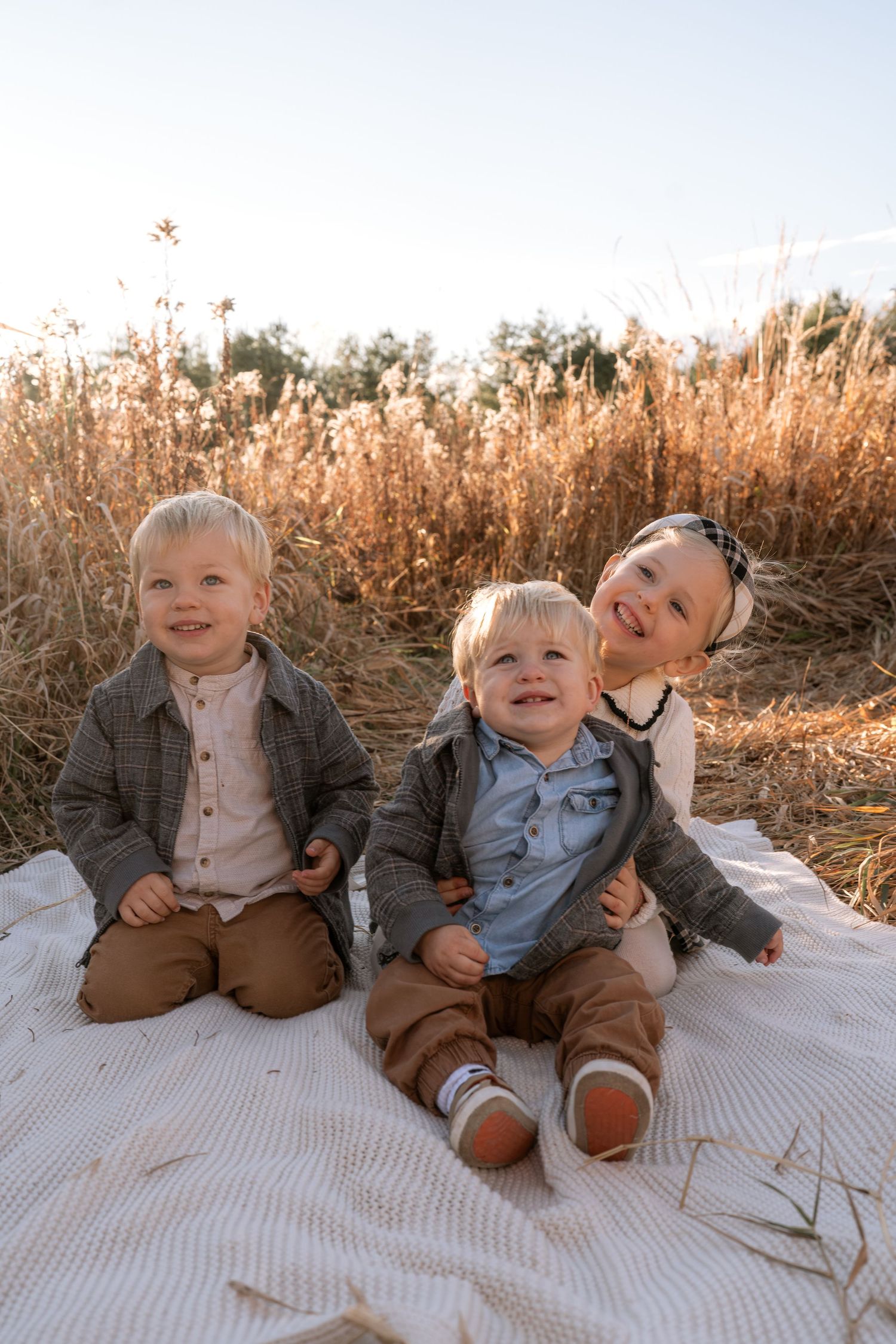 Young children sit together on white blanket in autumn field.