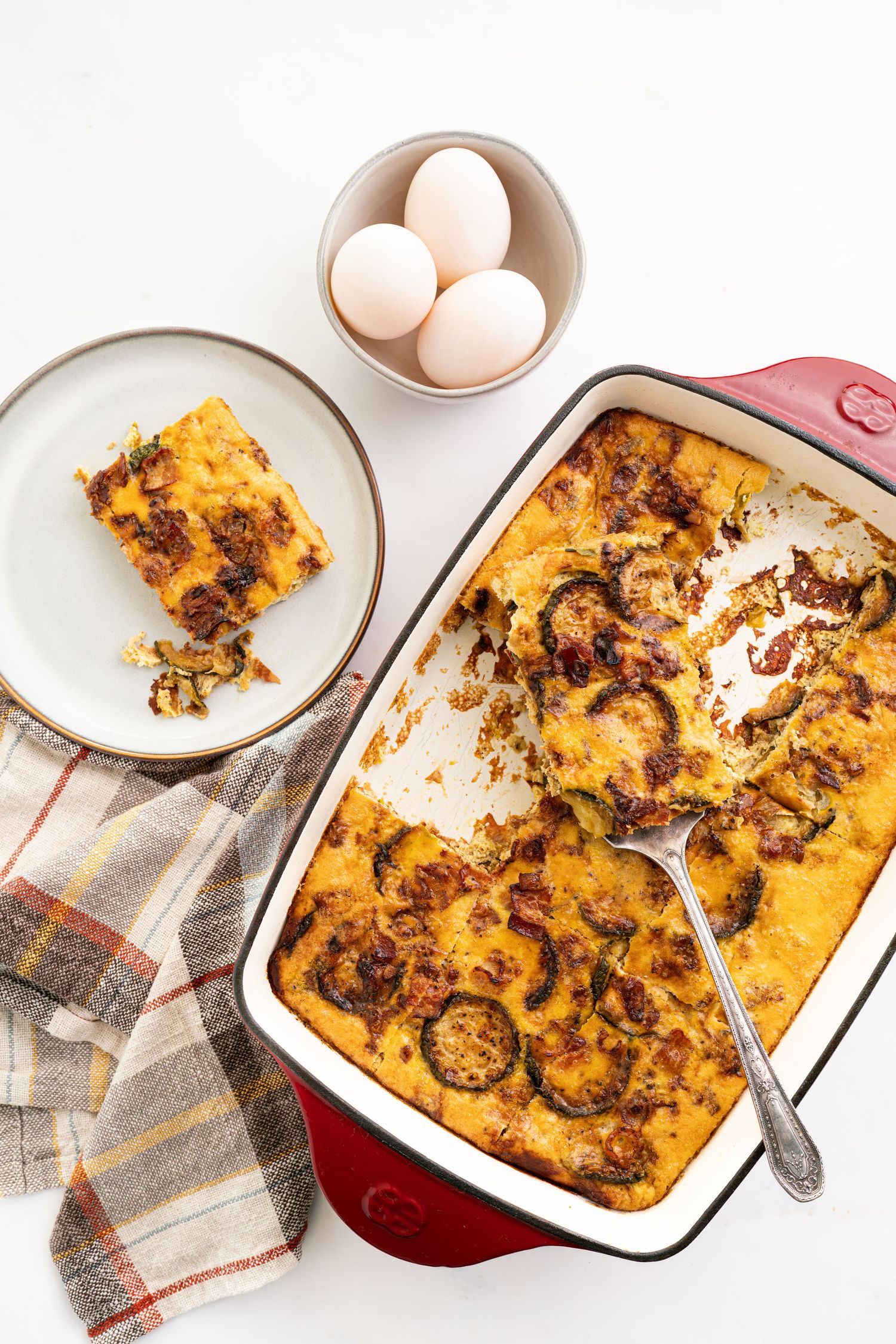 Breakfast casserole in red baking dish with a served portion and fresh eggs on the side.