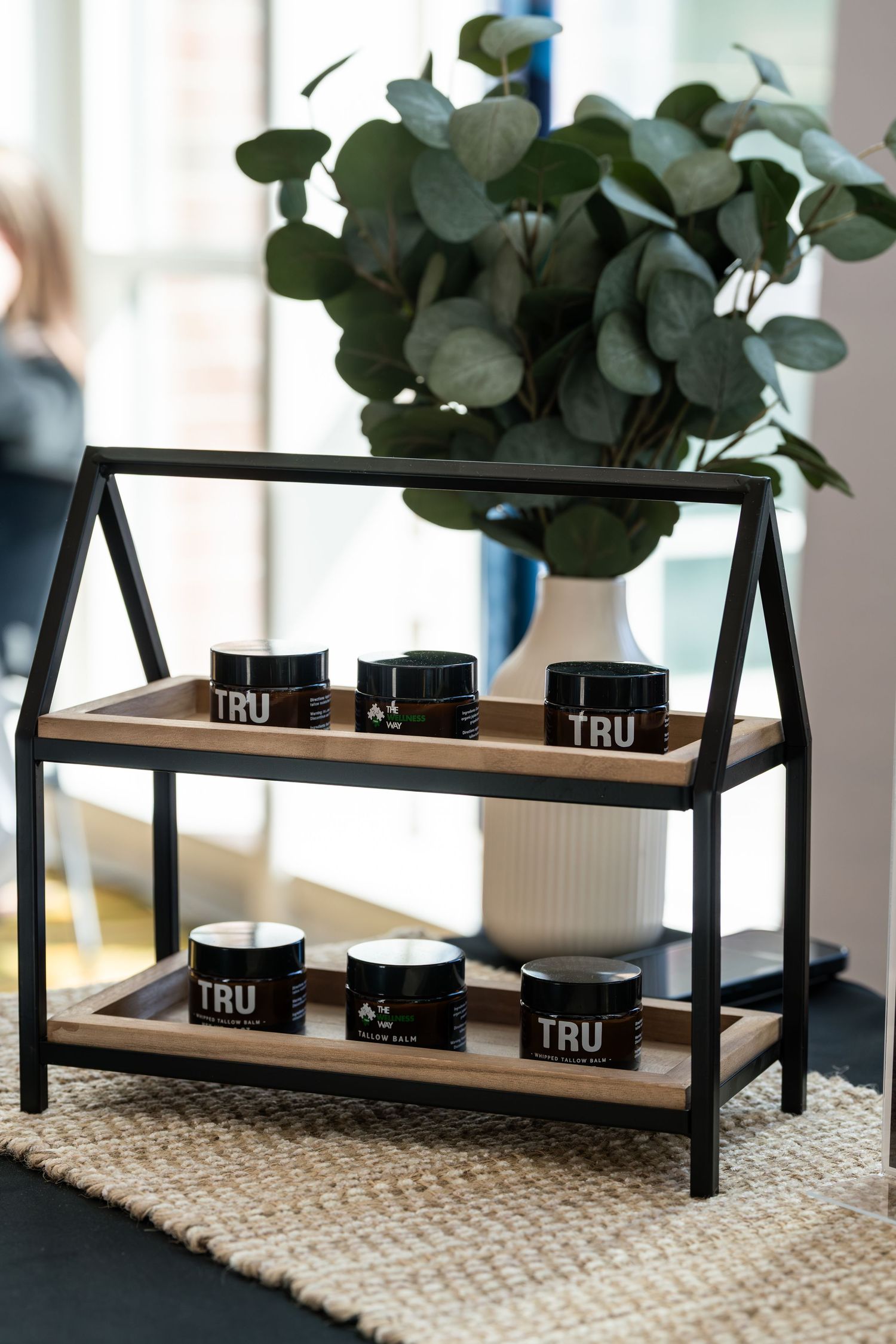 Modern black metal shelf display with TRU branded products and a potted eucalyptus plant on top.