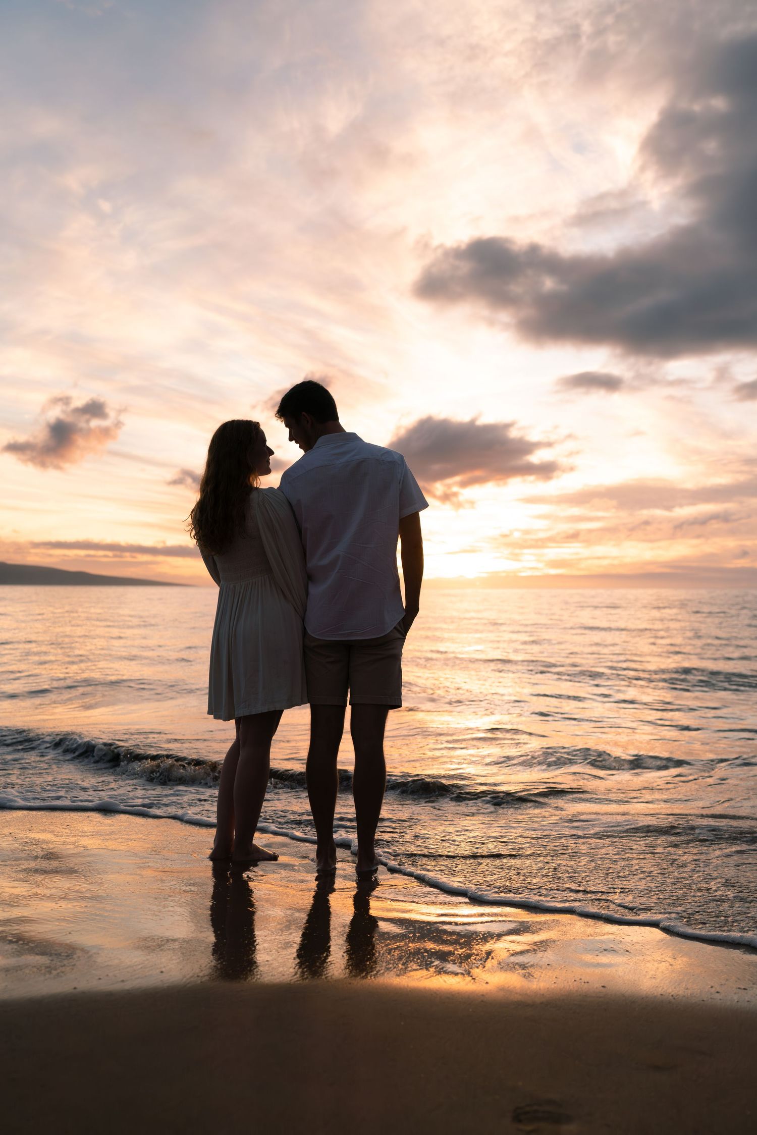 Silhouetted couple standing together on beach at sunset with waves lapping at their feet.