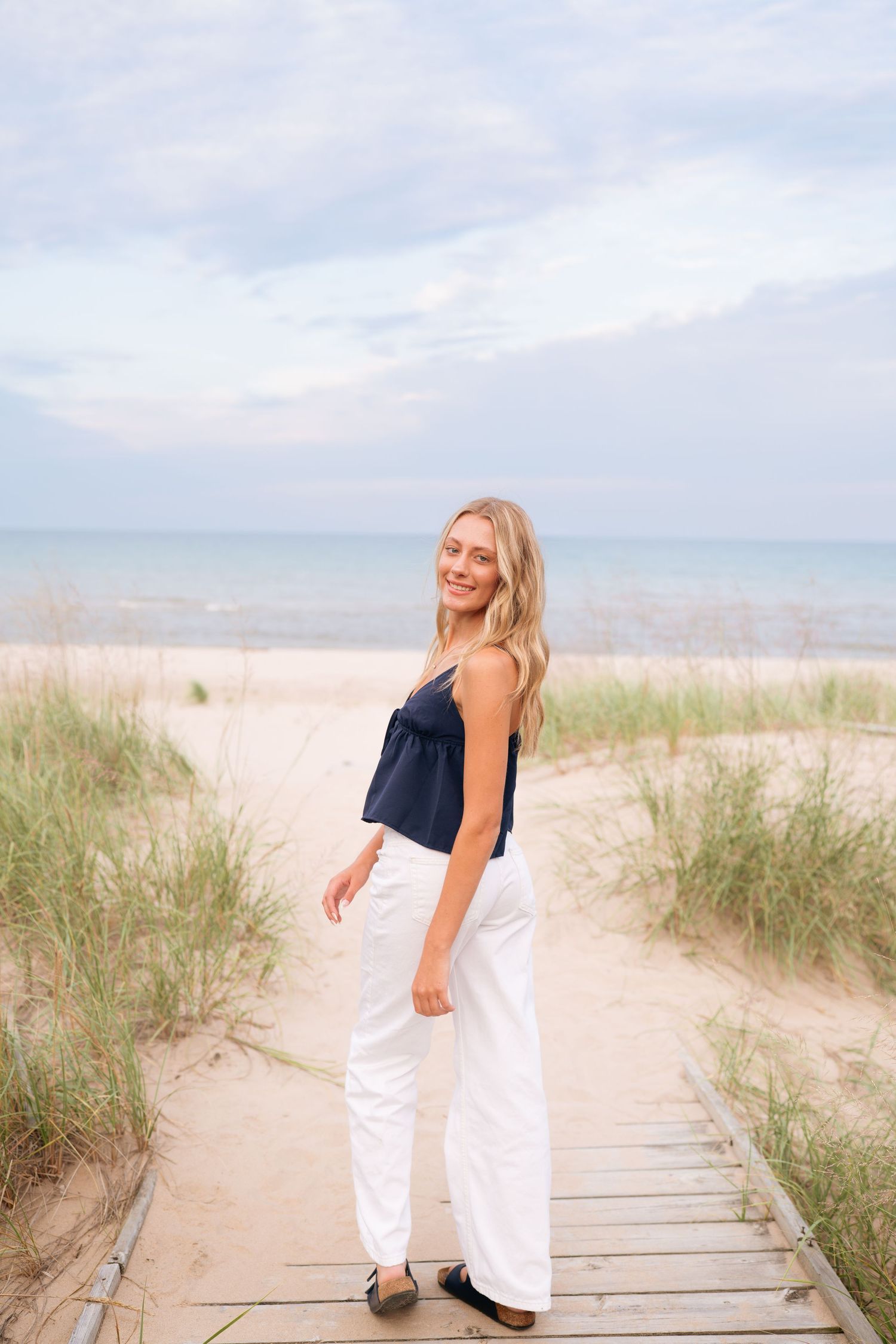 Woman in navy top and white pants poses on wooden boardwalk at beach during sunset.