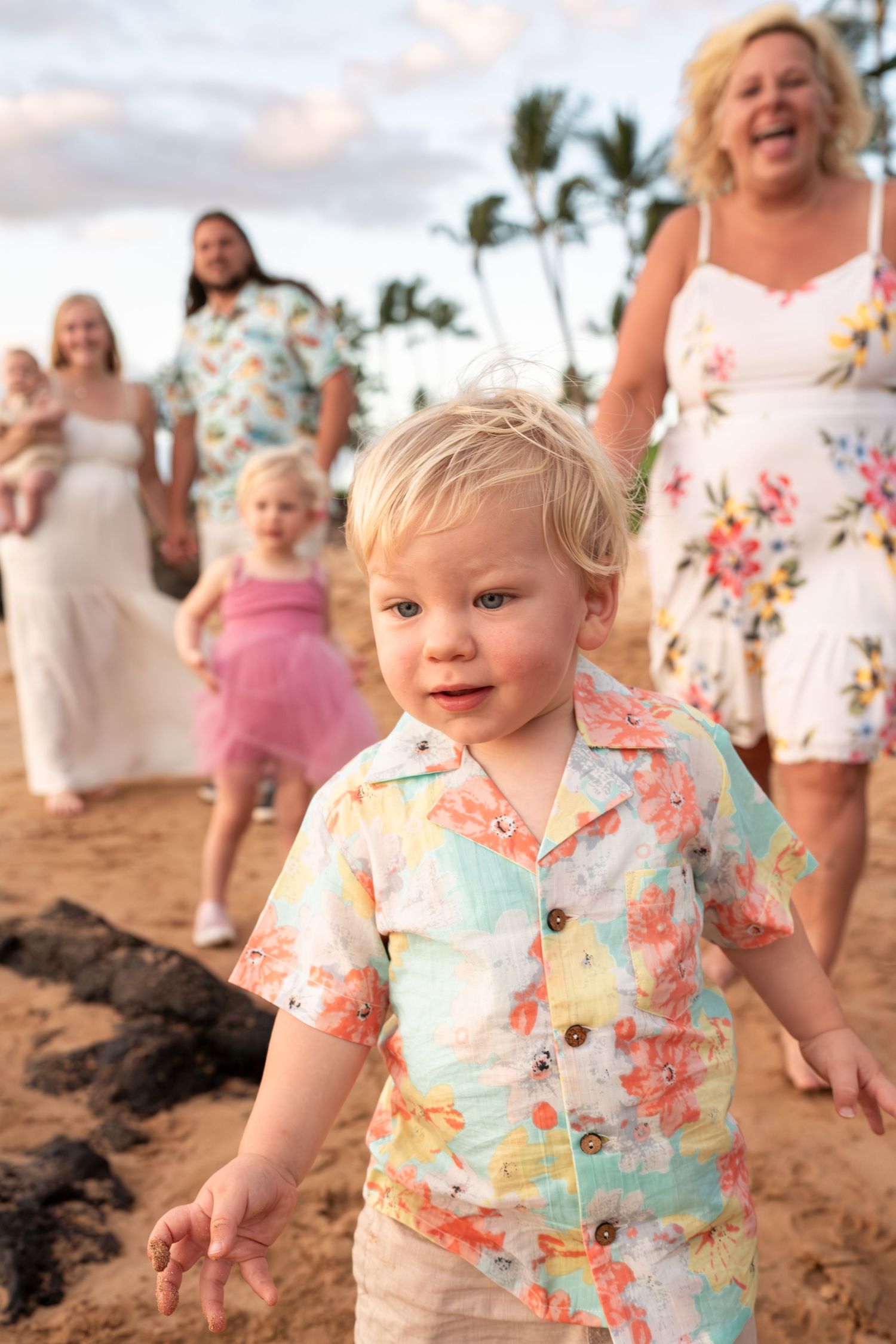 Toddler in floral shirt smiling and playing on sandy beach at sunset.