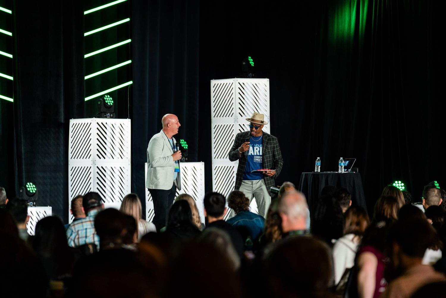 A presenter speaks on stage with green lighting and geometric backdrop at a professional conference.