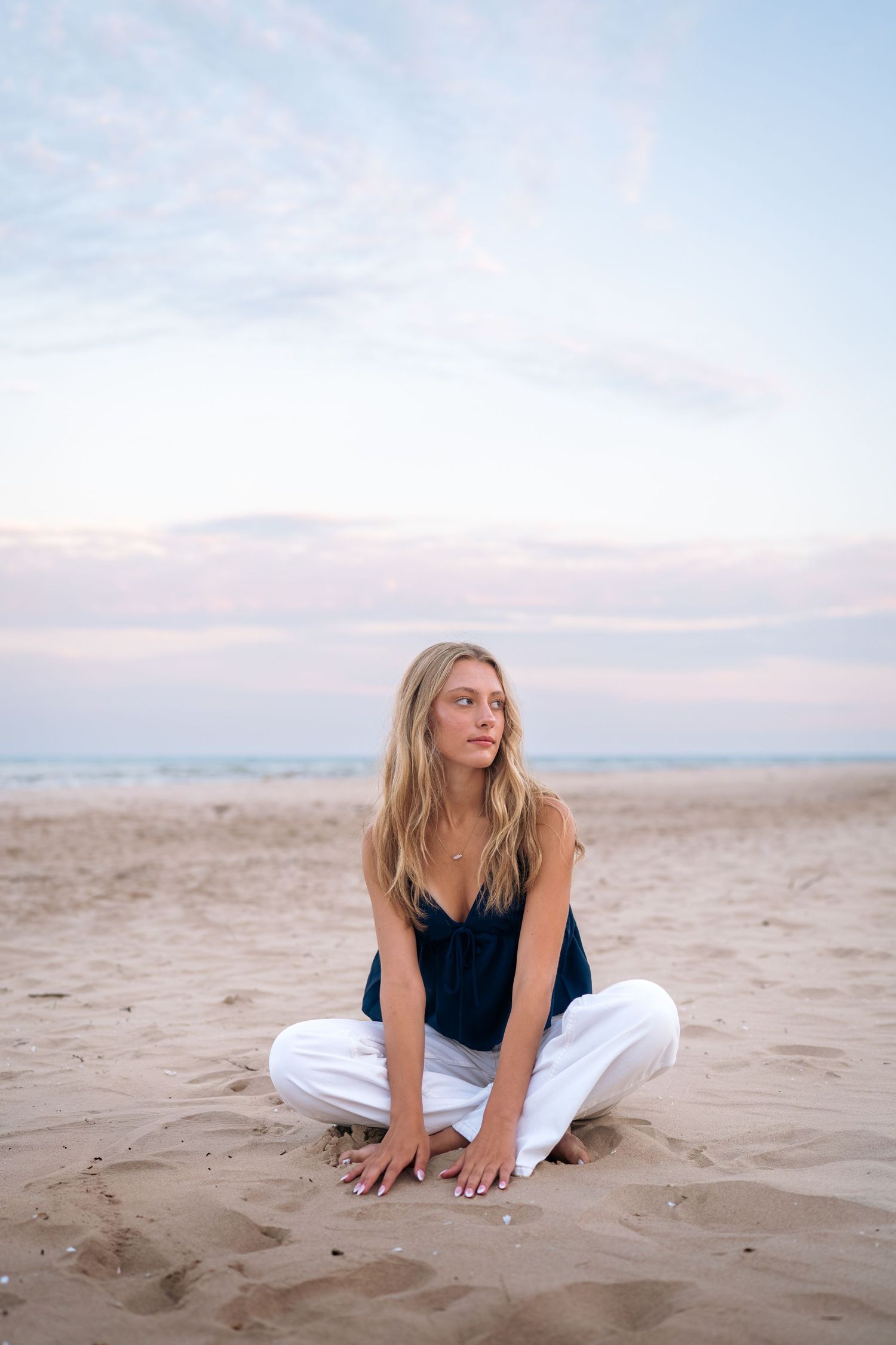 Woman in black top and white pants relaxes on sandy beach at dusk.