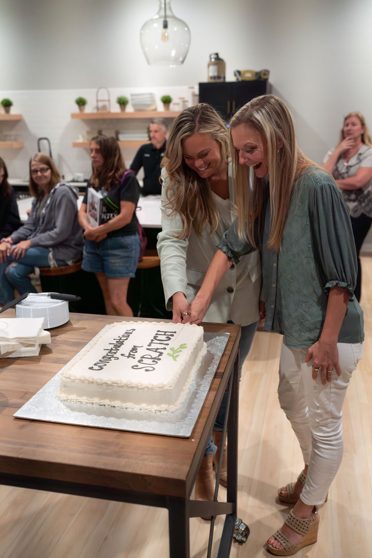 A celebration cake sits on a wooden table while people gather around to cut it.