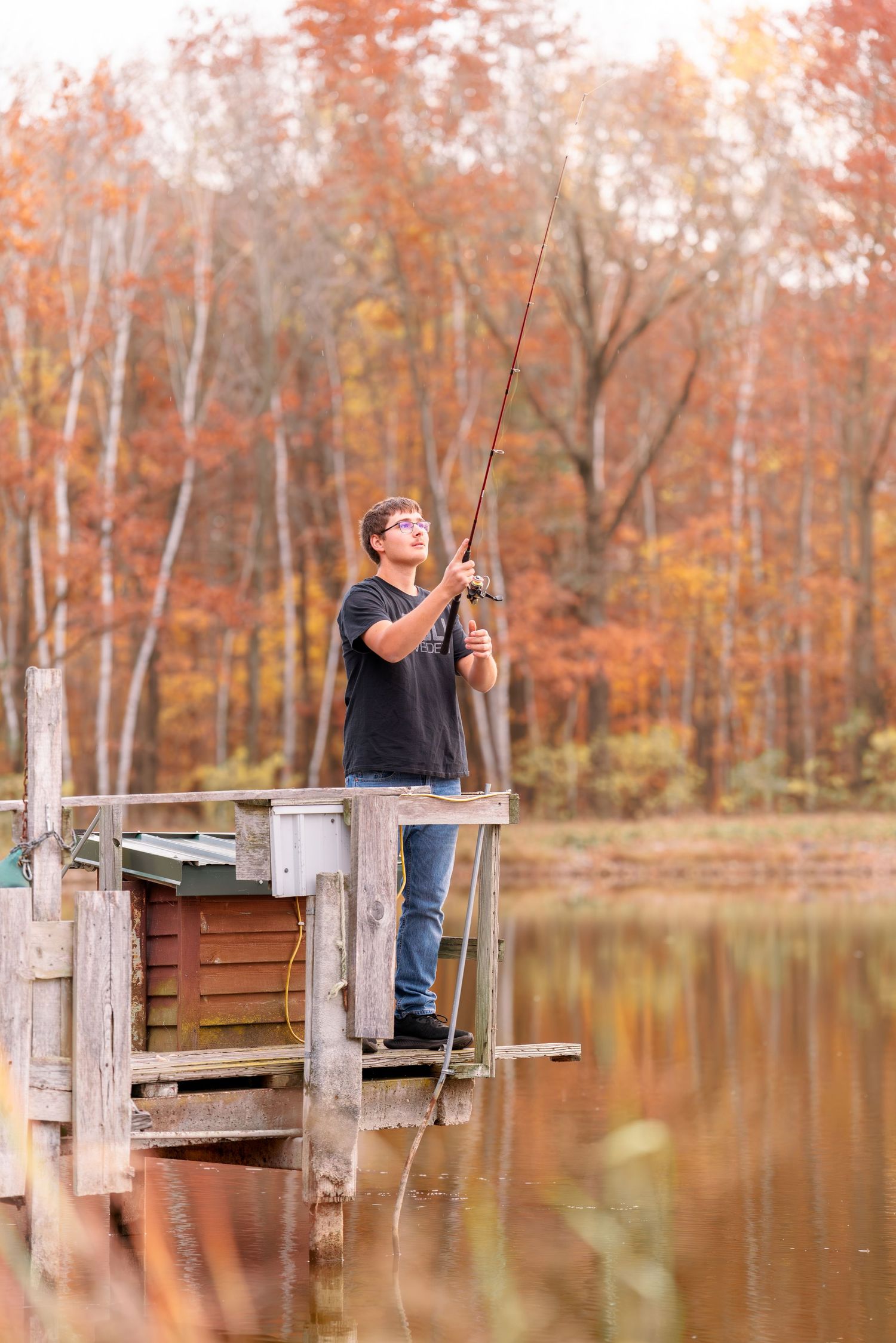 Person fishing from a wooden dock on a tranquil lake surrounded by autumn foliage.