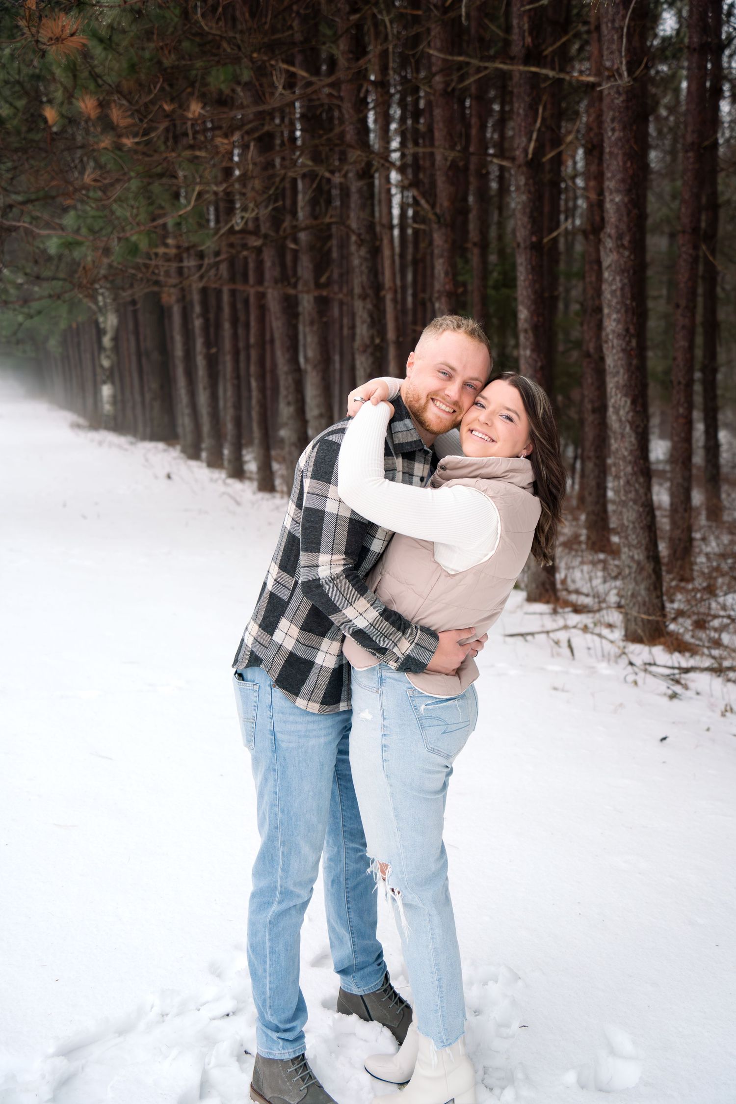 Couple embracing in snowy winter forest with pine trees in background.