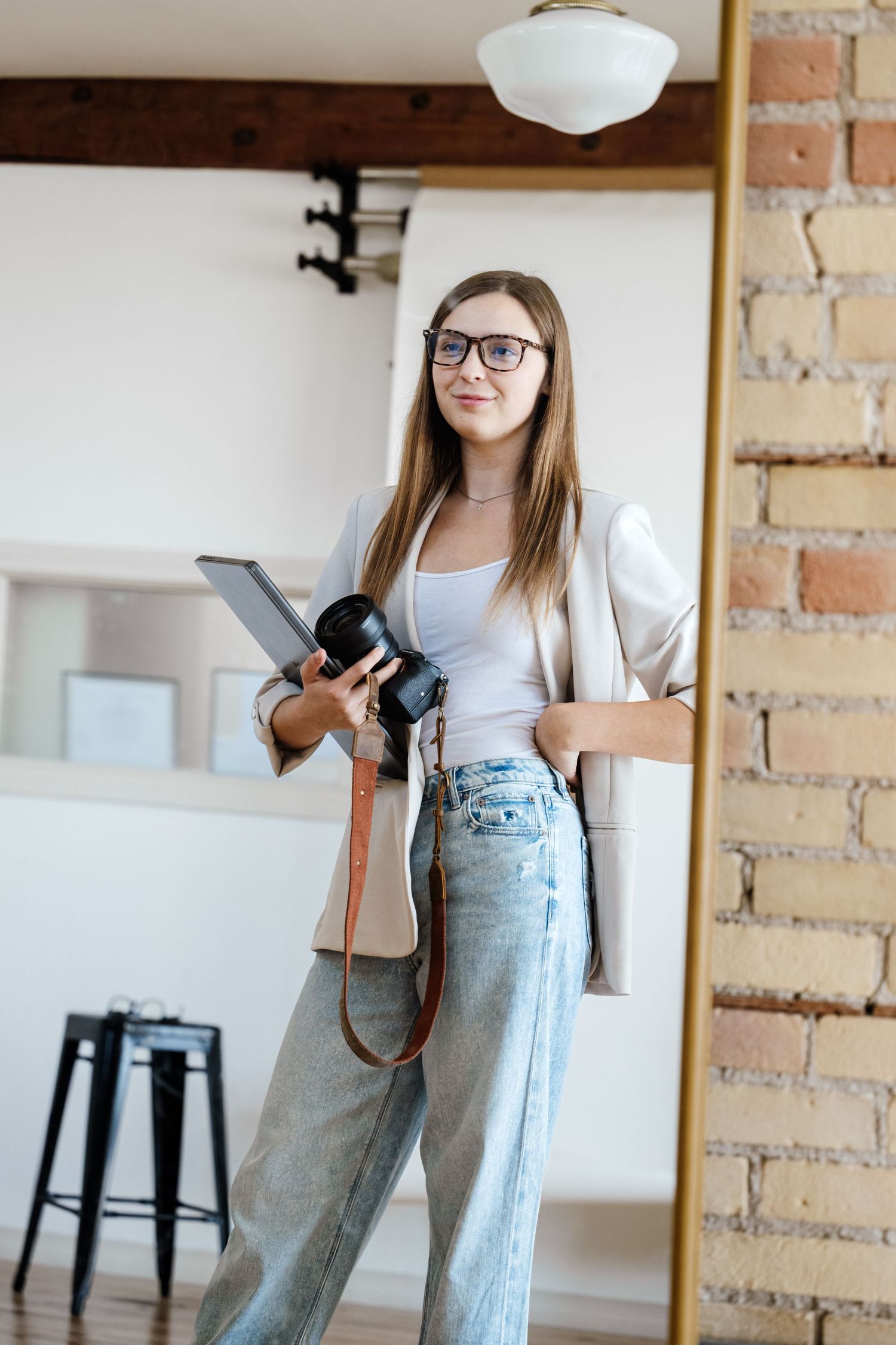 Photographer in casual attire holding camera in studio with exposed brick wall.