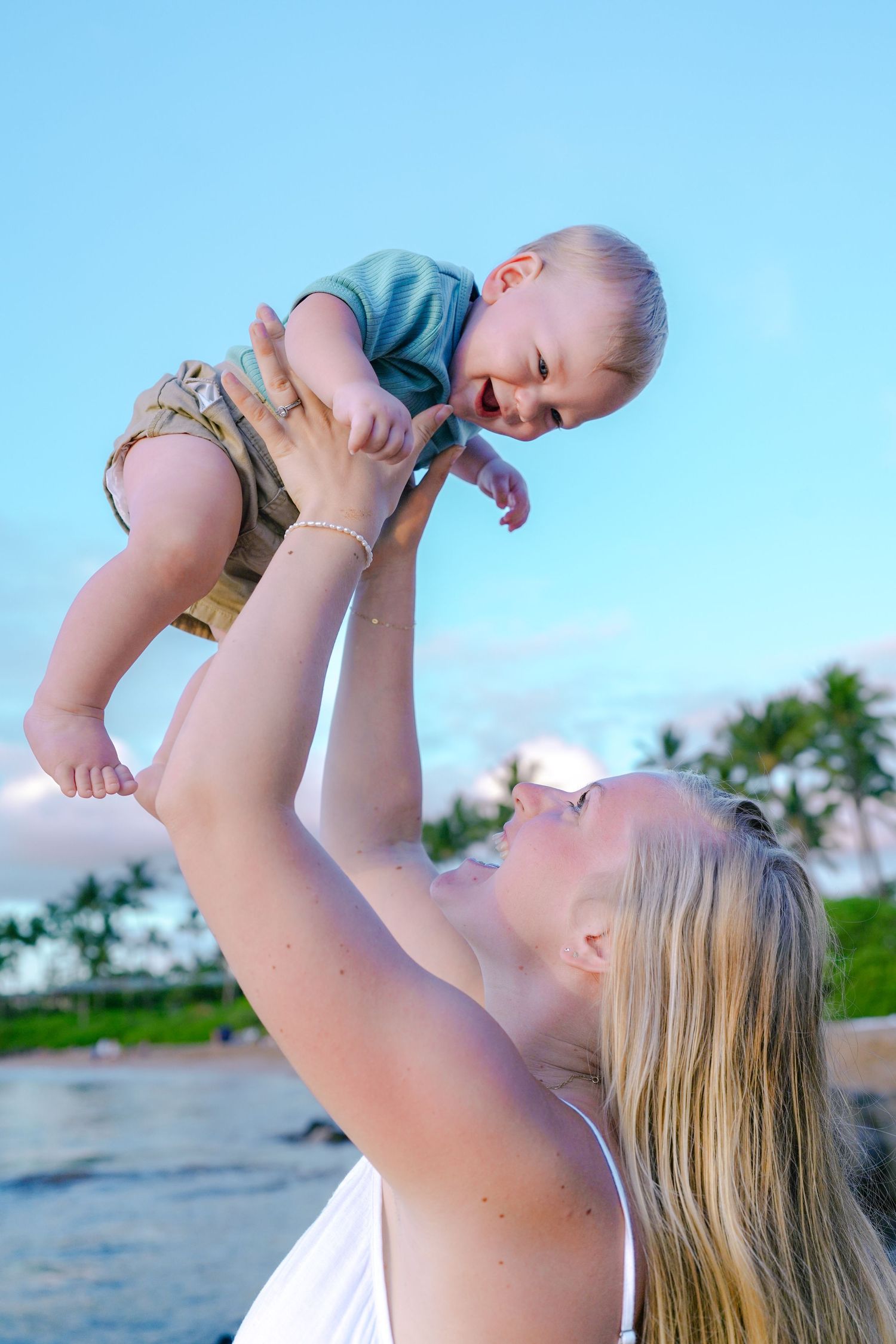 Parent lifting baby up against blue sky at beach during sunset.