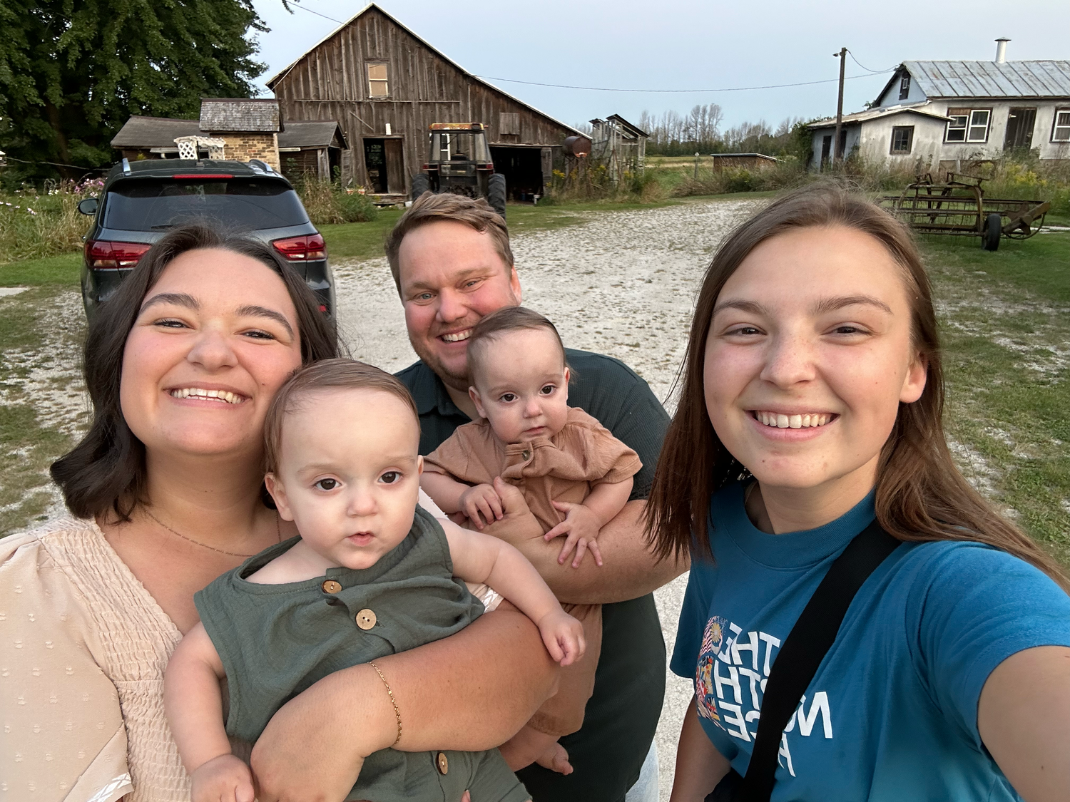 A group takes a selfie together with infants in front of a rustic barn on a country road.