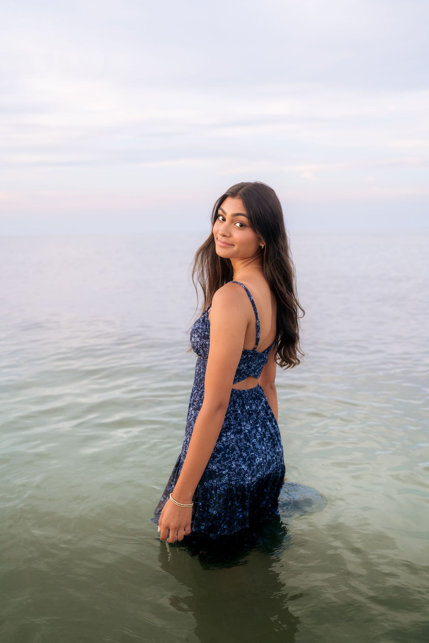 A person stands in ocean water wearing a navy floral dress during sunset.