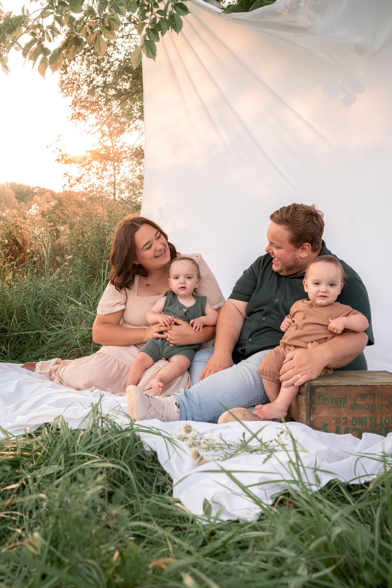Family lying together on white blanket in grassy field during golden hour sunset.