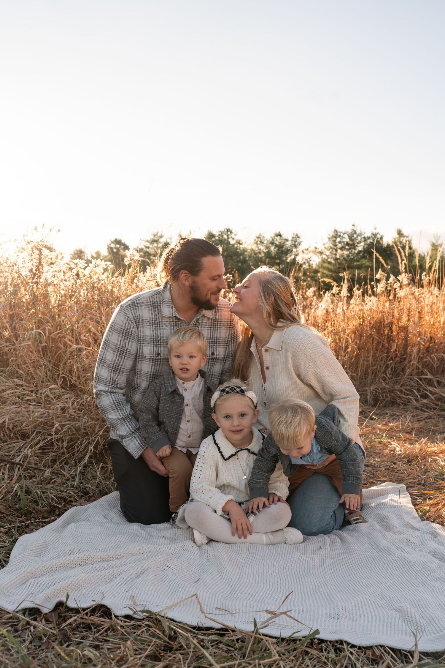 Family sits together on white blanket in golden field during sunset.