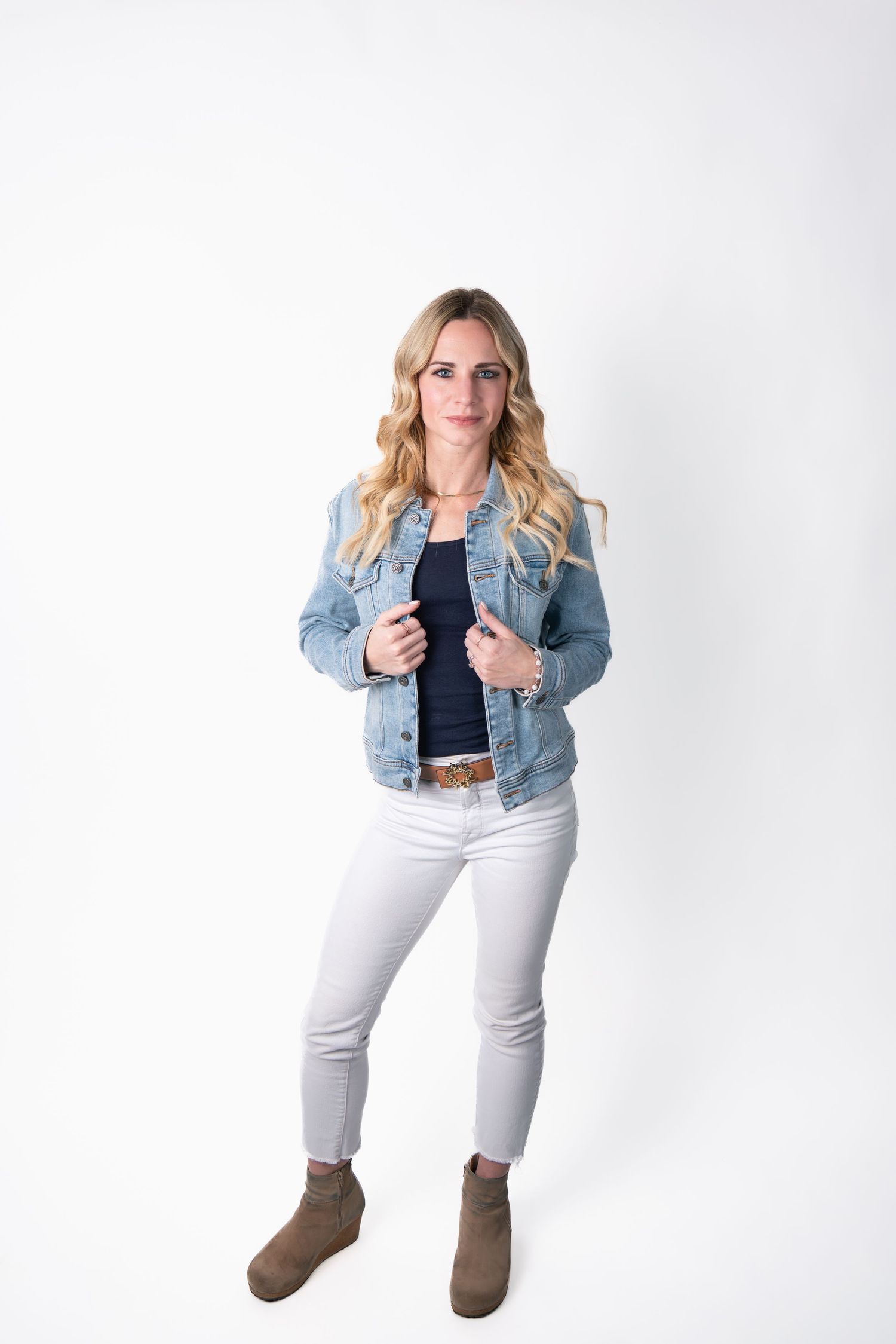 Model wearing denim jacket and white pants poses casually against white studio backdrop.