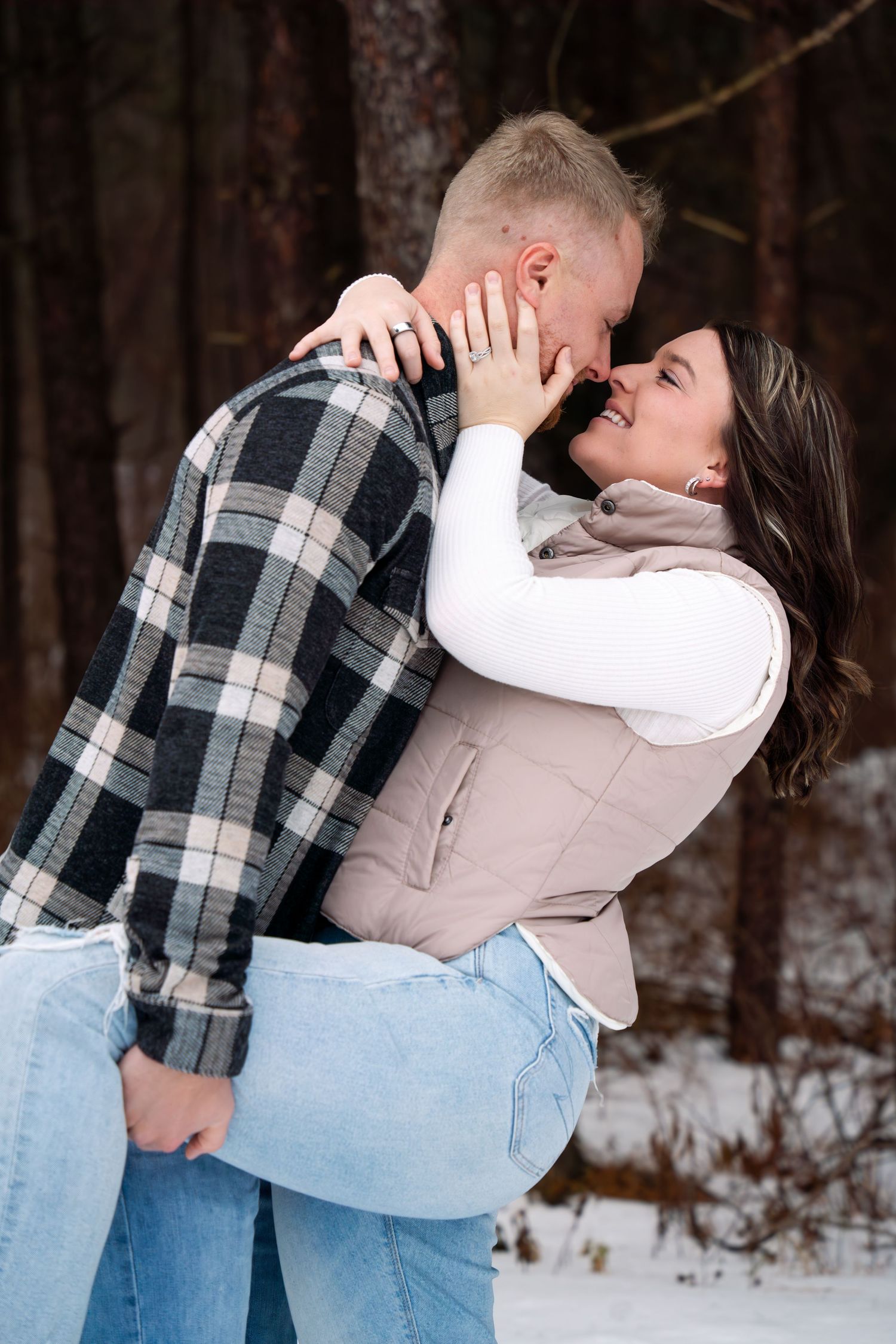 Couple embracing in romantic winter photoshoot with snow-covered trees in background.