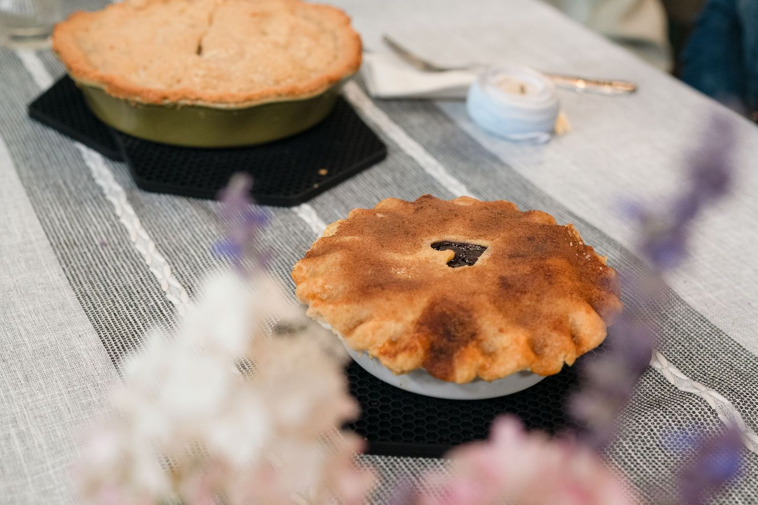 Two freshly baked golden brown pies with decorative crusts sit on a table with blurred flowers in the foreground.