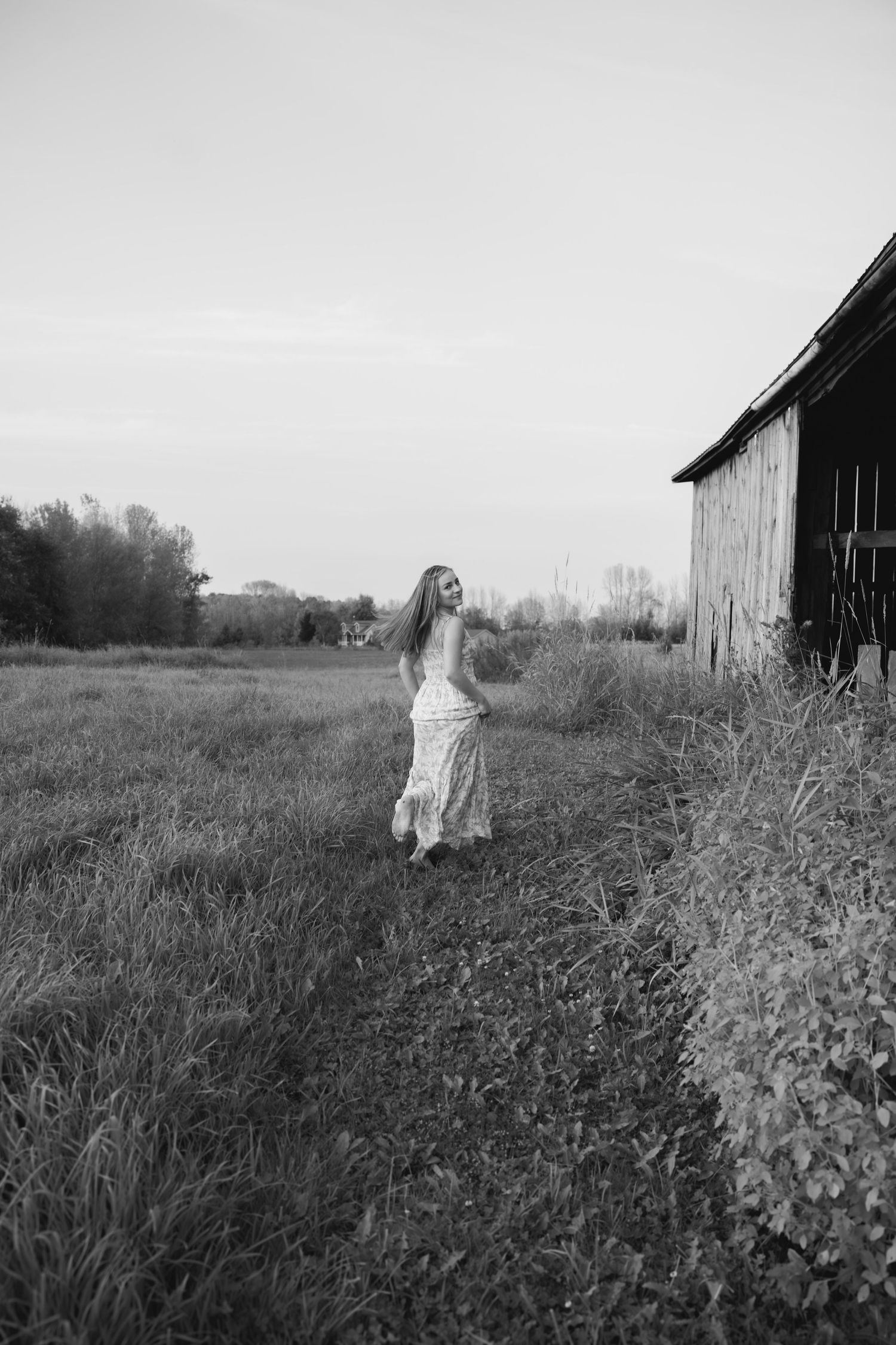 Black and white artistic photo of ethereal figure near rustic barns in tall grass.