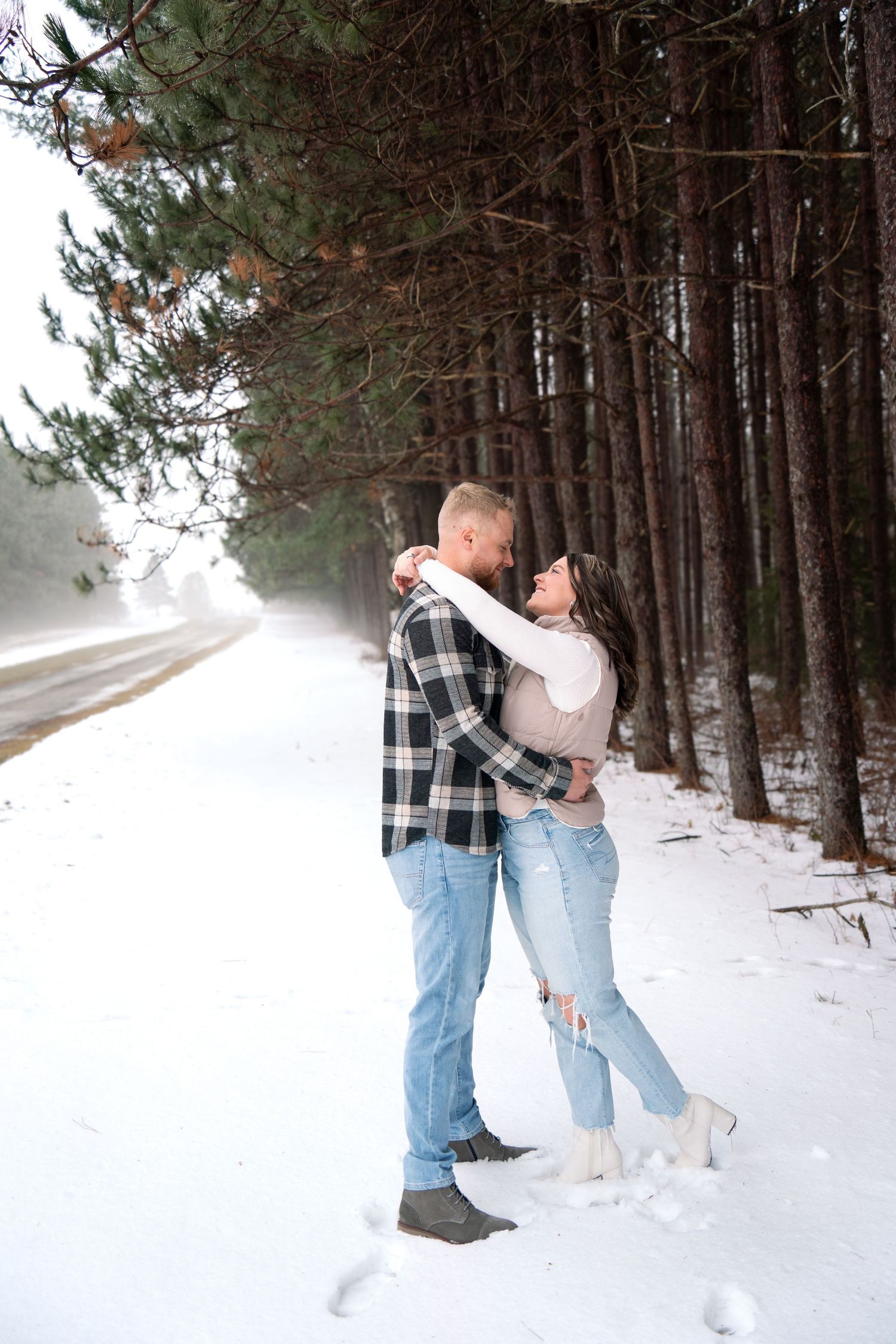 Playful couple posing for photos on snowy winter path through pine forest.