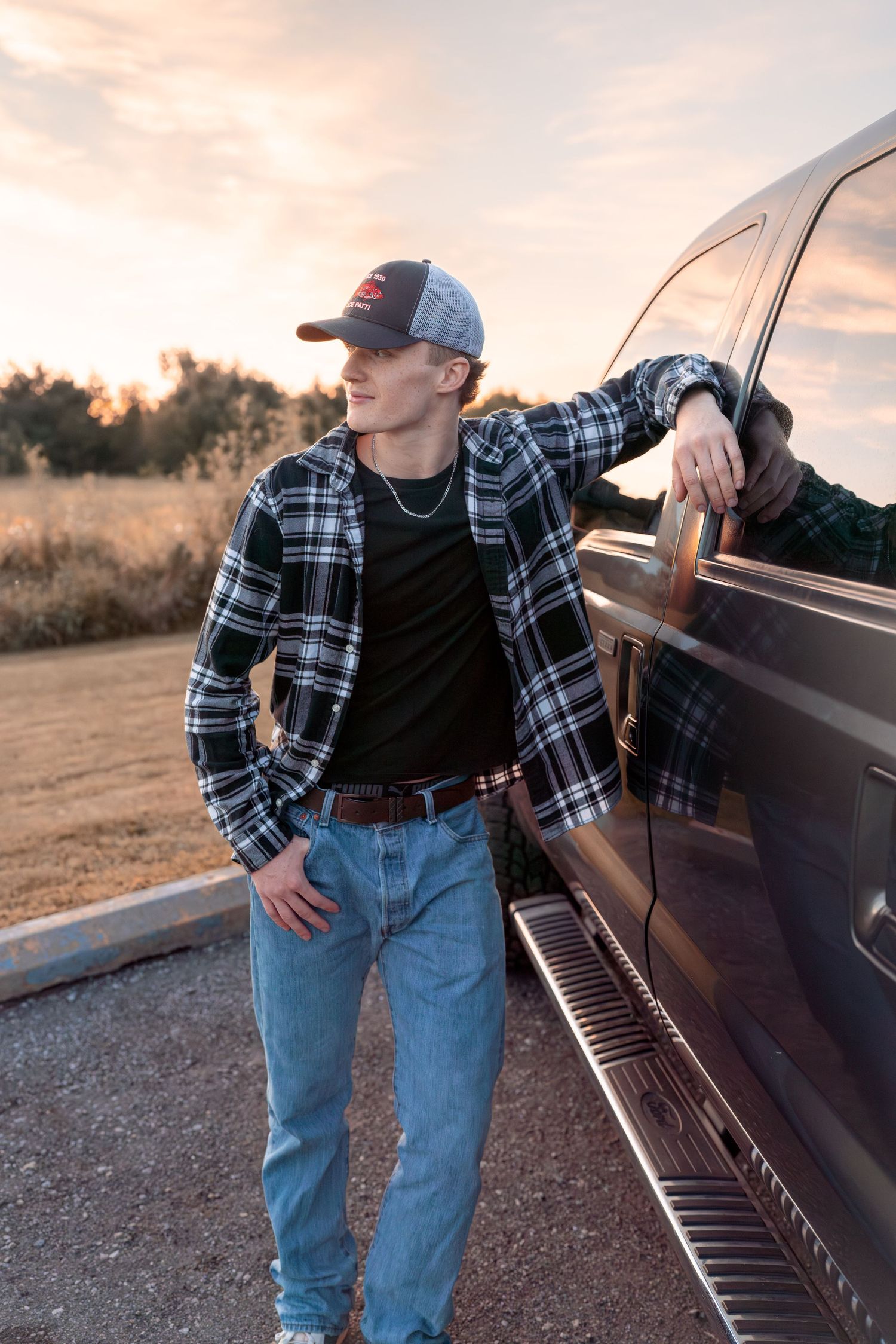 Person in plaid shirt and jeans leans against vintage car at sunset.