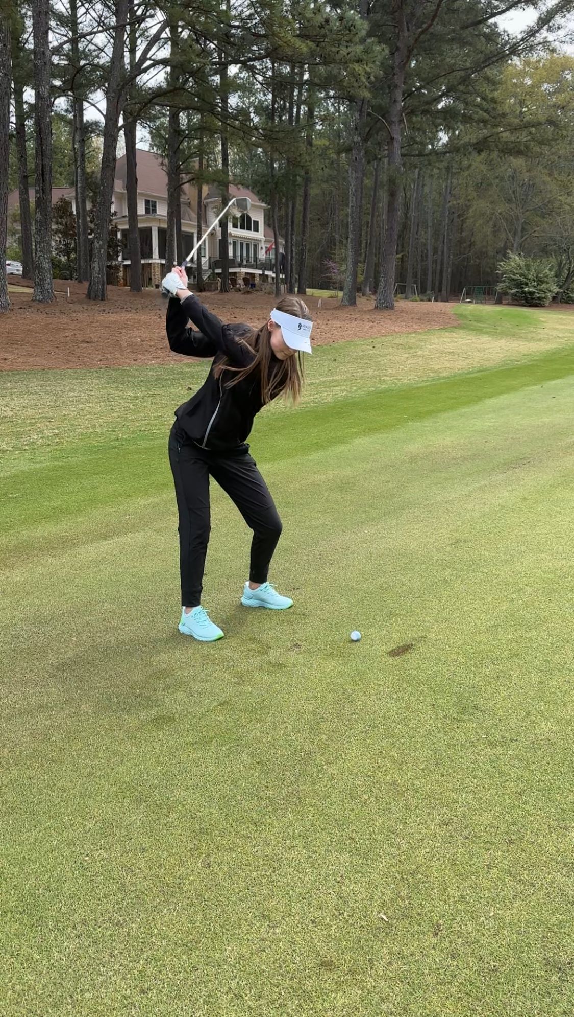 A golfer in black athletic wear takes a swing on a scenic course surrounded by pine trees.