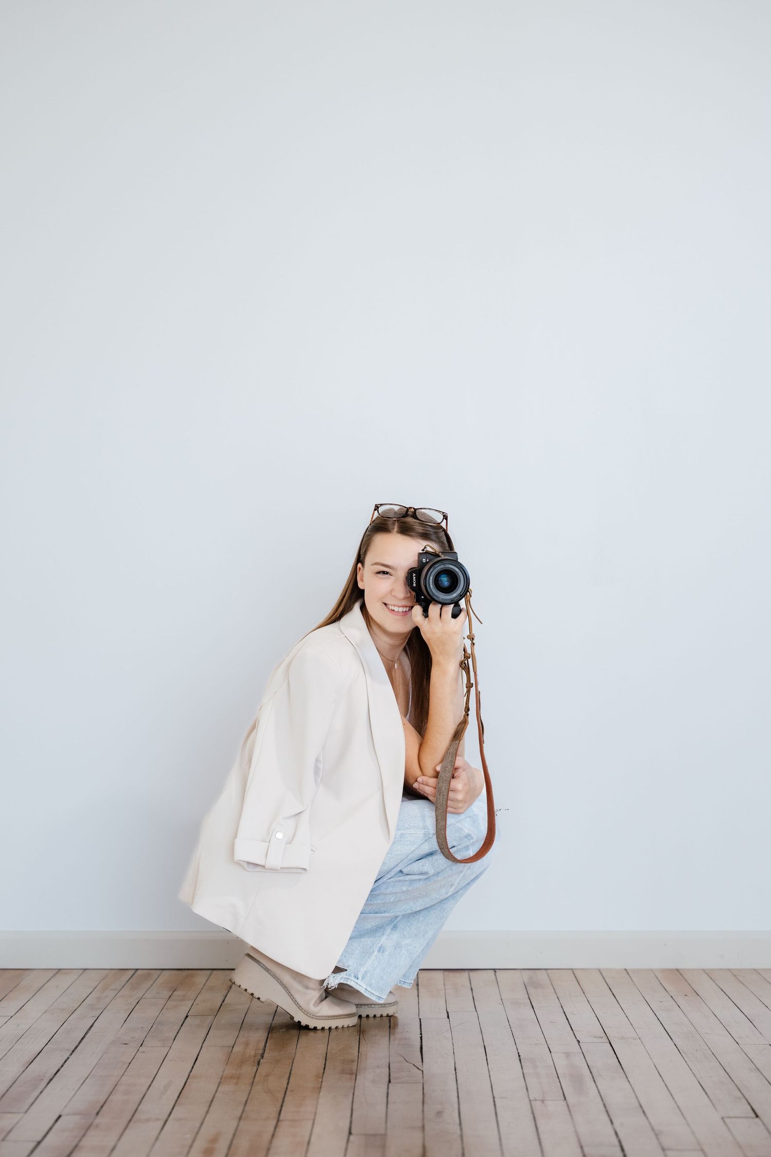 Photographer in white fabric crouching on wooden floor with camera against minimal white backdrop.