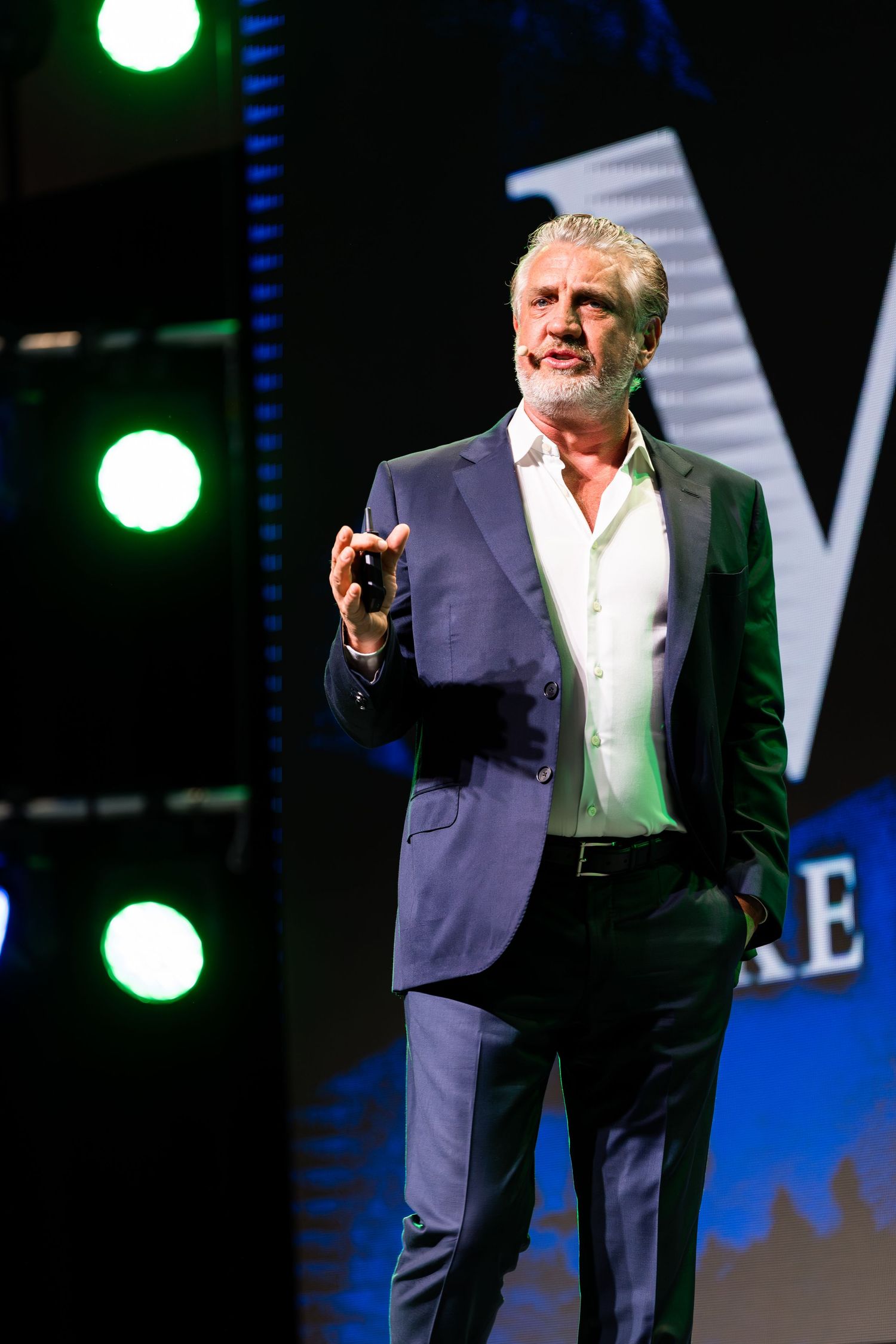 A speaker in a navy suit gestures energetically on stage with dramatic lighting effects.