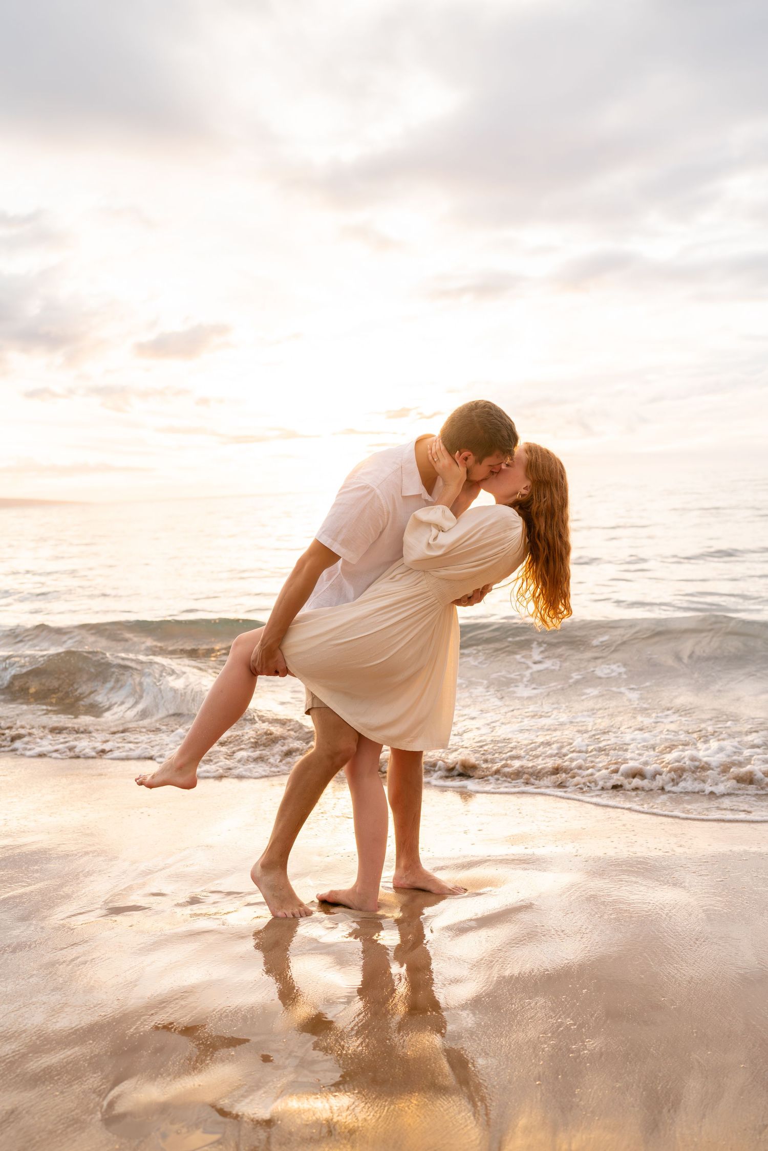 Romantic beach scene with couple in white attire playing in ocean waves at sunset.