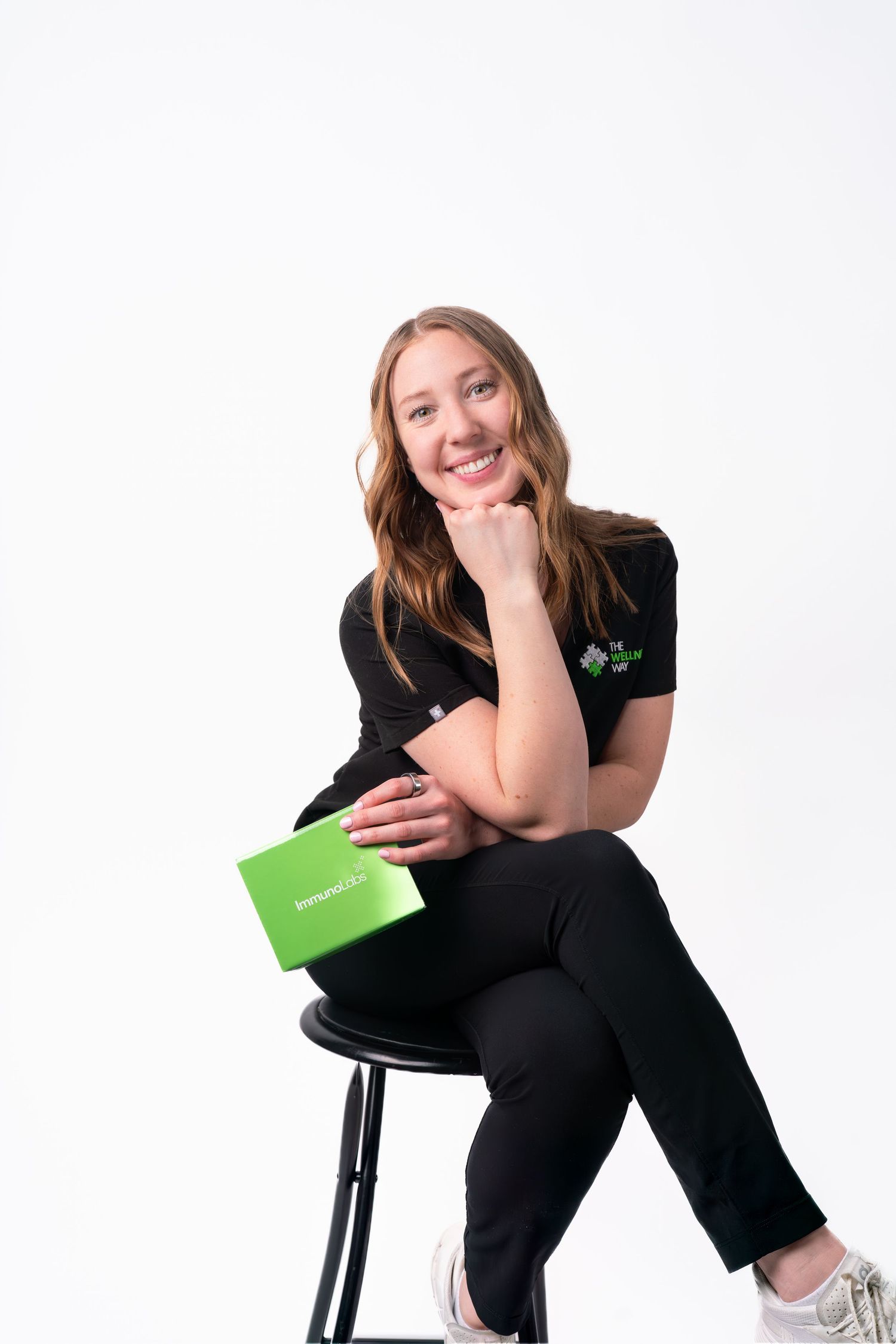 Person in black clothing sits casually on a chair with a green book against white background.