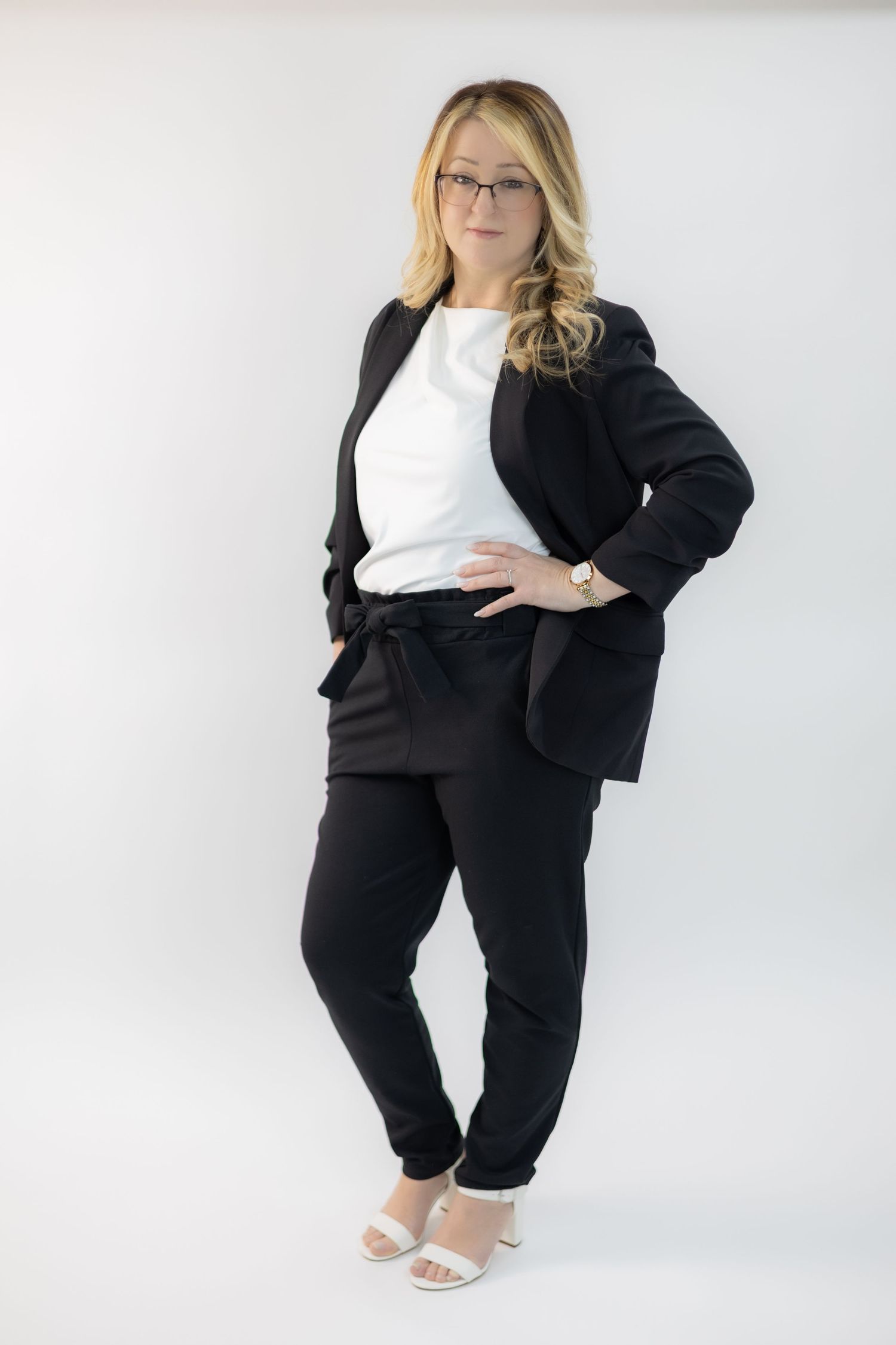 Professional woman in black blazer, trousers, white top, and heels poses confidently against a white background.