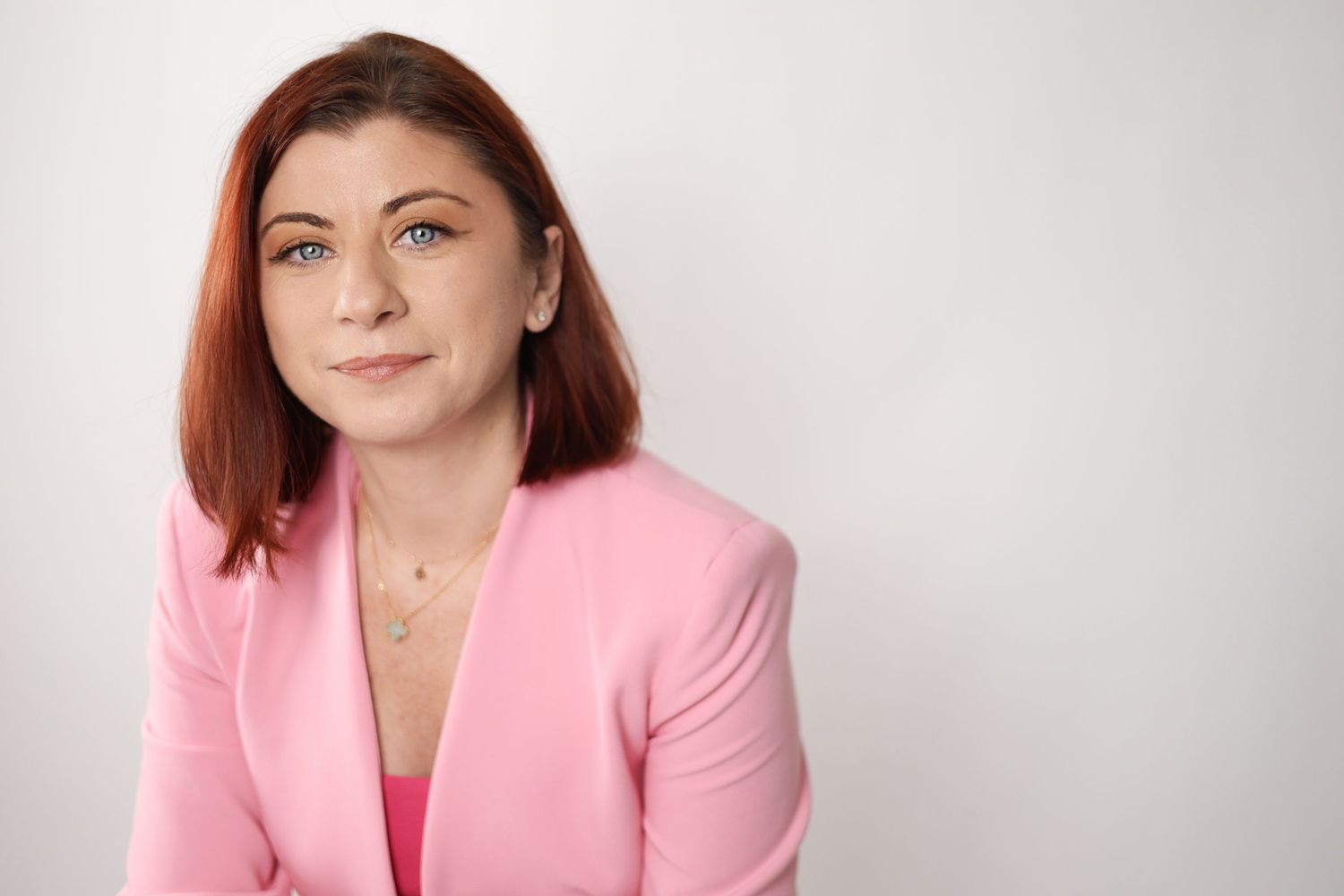 Professional woman with red hair wearing a pink blazer posing against a white background.