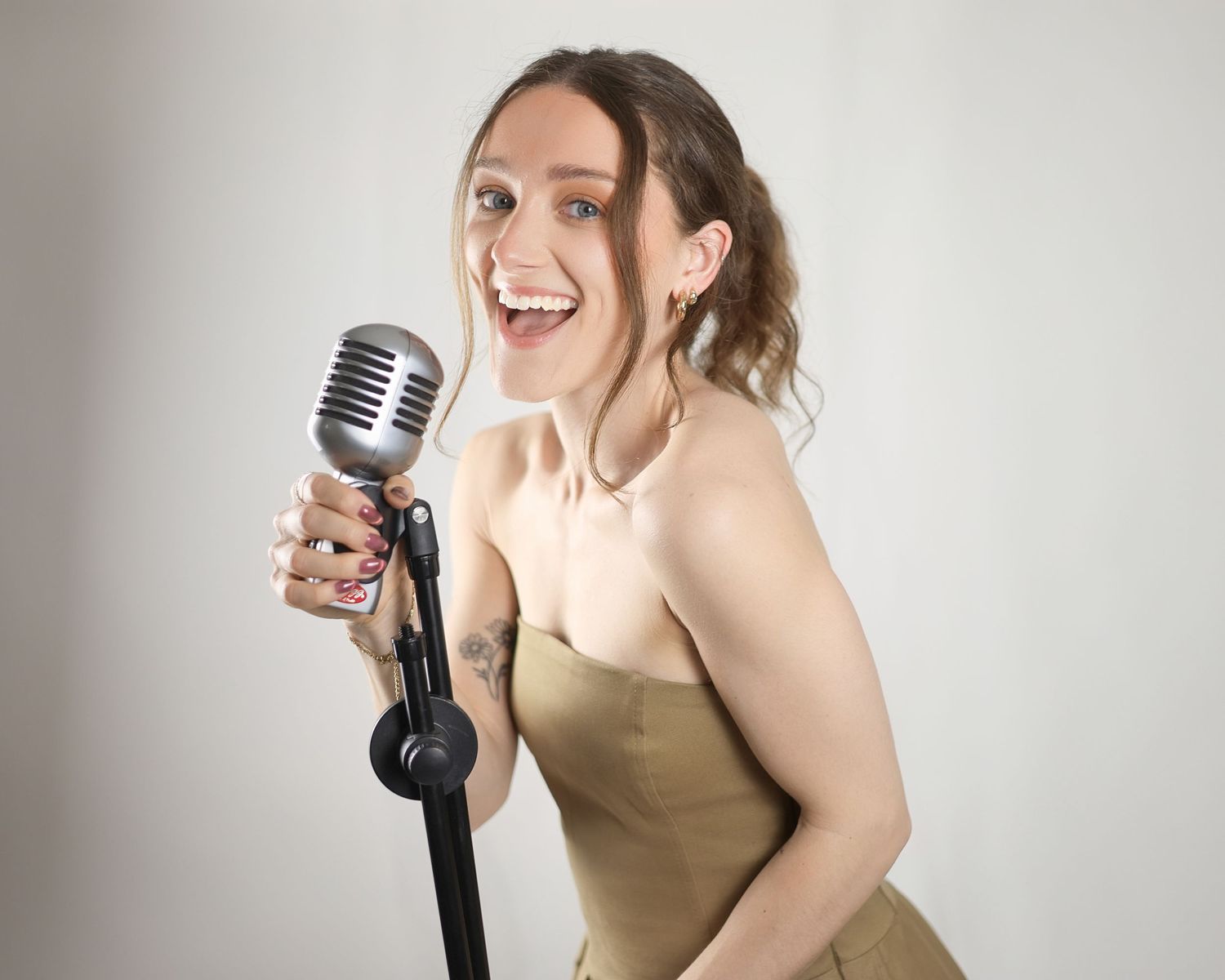 Young woman in gold strapless dress singing joyfully into a vintage silver microphone on a stand.