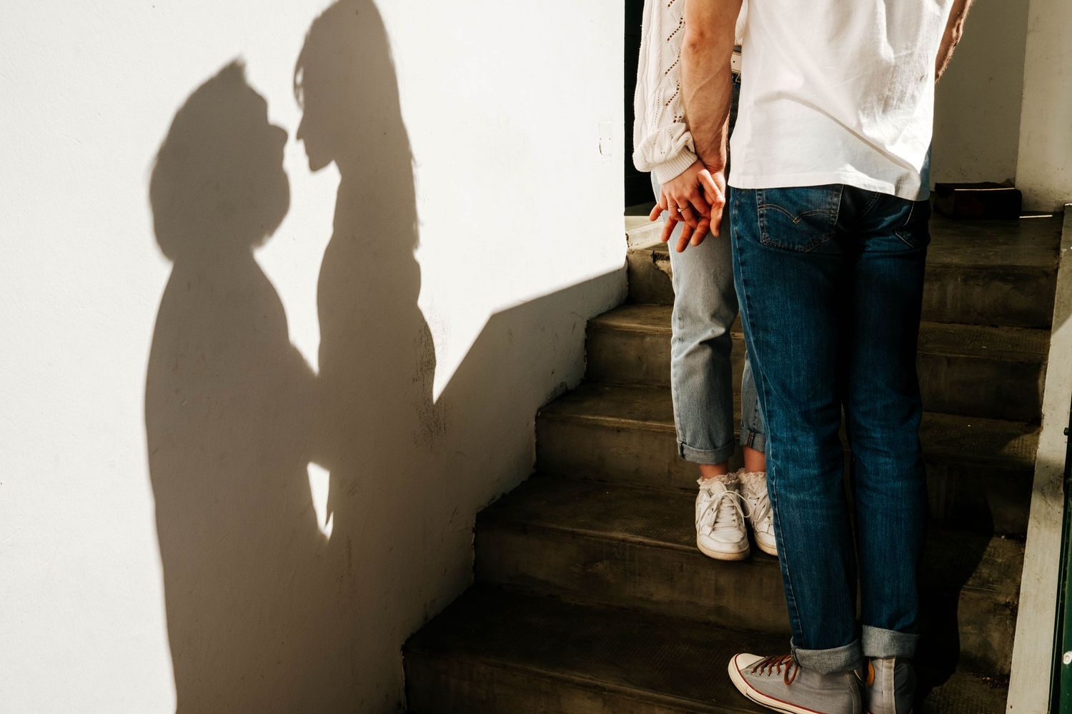 Couple kissing on stairs with romantic shadow silhouette on white wall, wearing jeans and white tops.